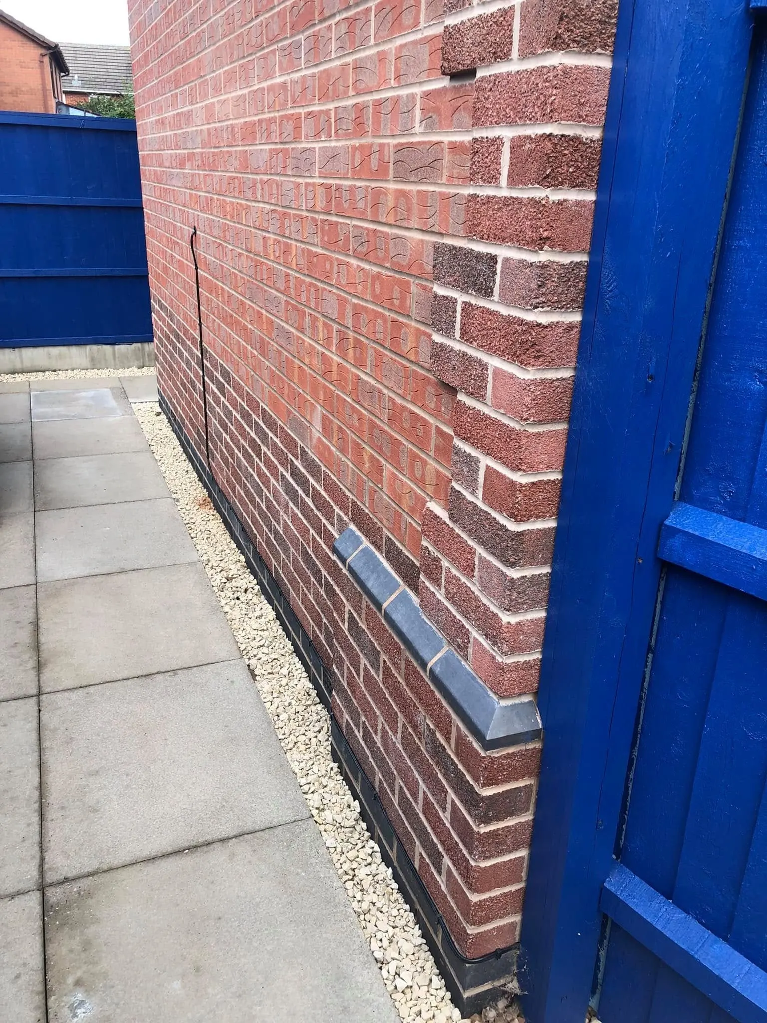 Black cable clipped down a red brick wall and along the dark base course of a side passage beside a blue fence