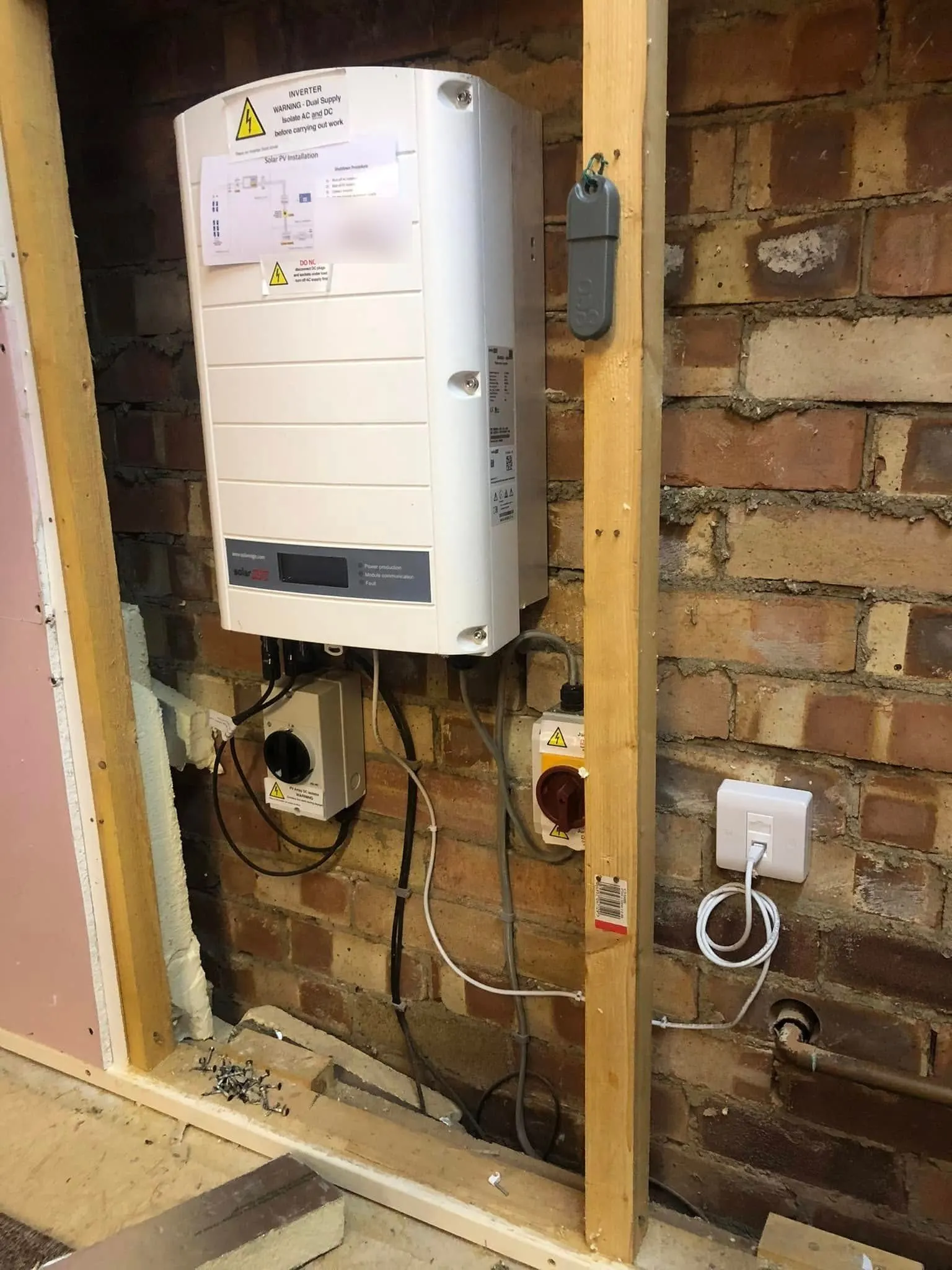 White ethernet wall socket with a coiled patch lead on a brick wall beside a SolarEdge inverter and isolator switches