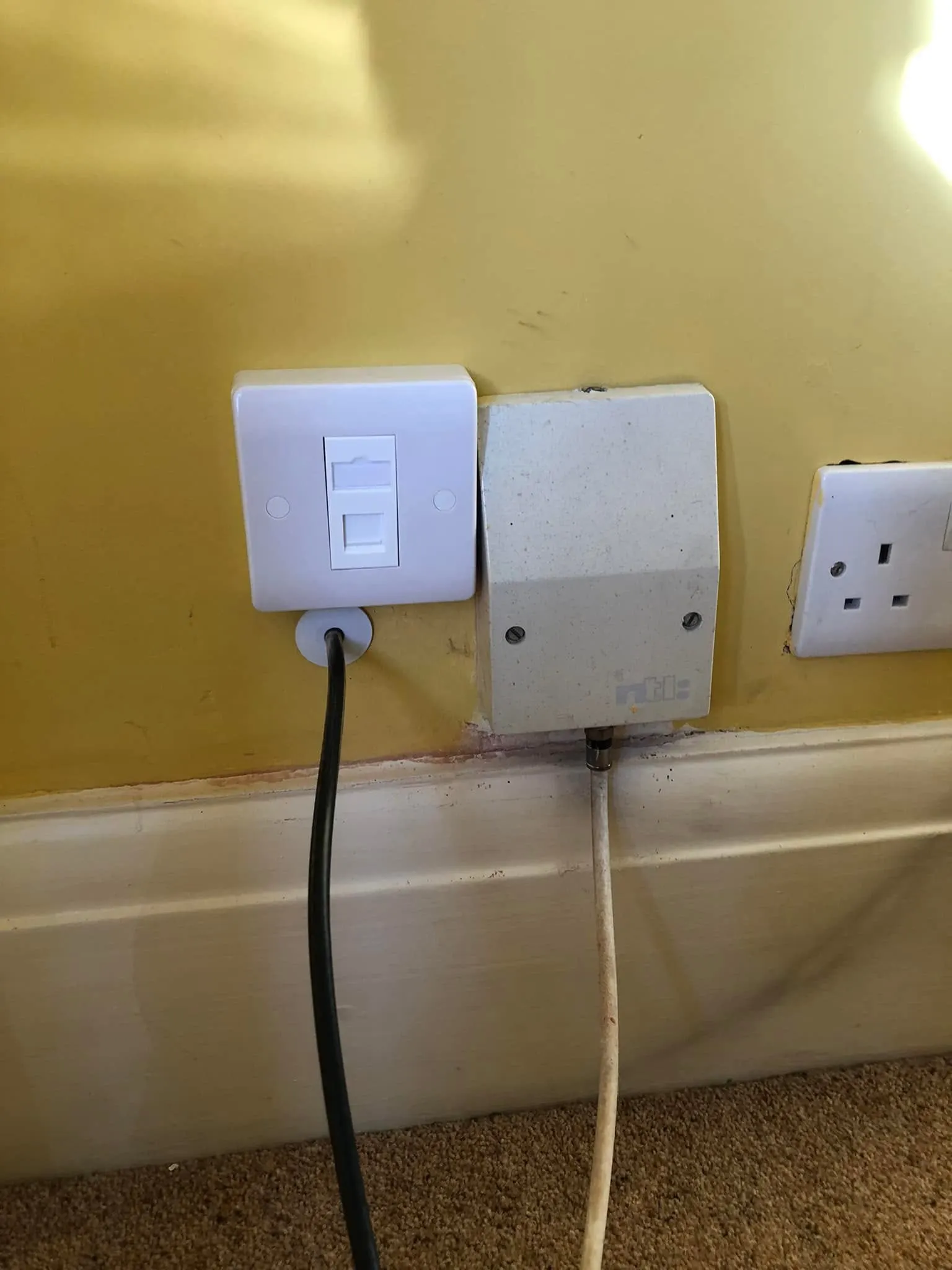 White single ethernet faceplate on a yellow wall beside an old NTL box, with a black cable running down from a grommet below it