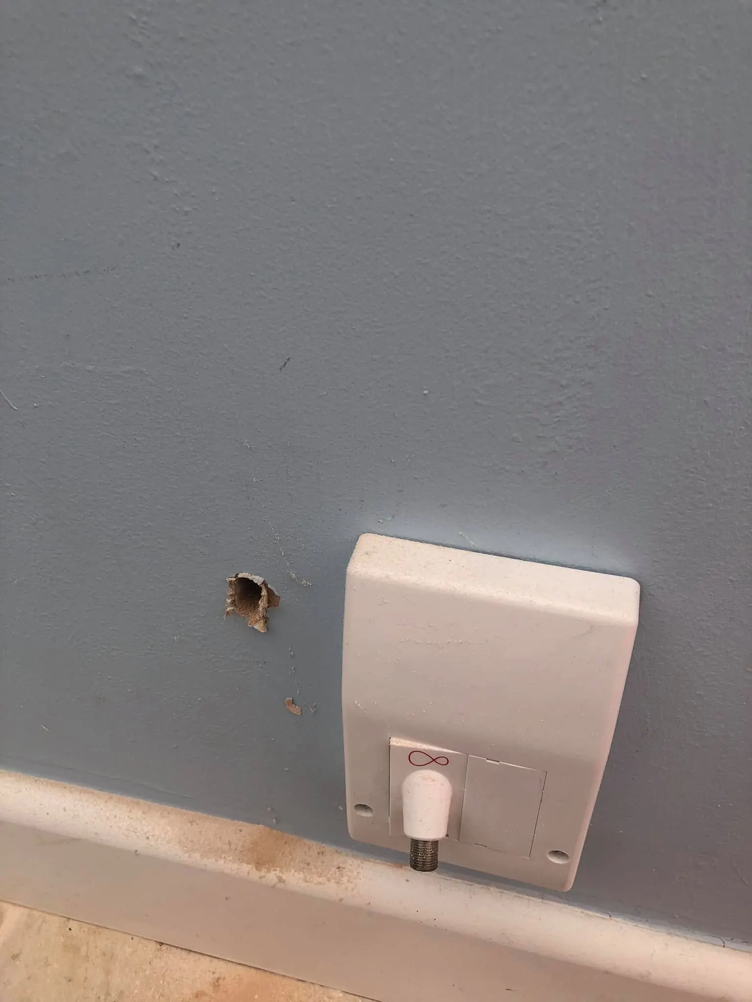 Rough drilled hole in a pale blue wall beside a white Virgin Media coax wall box above the skirting board