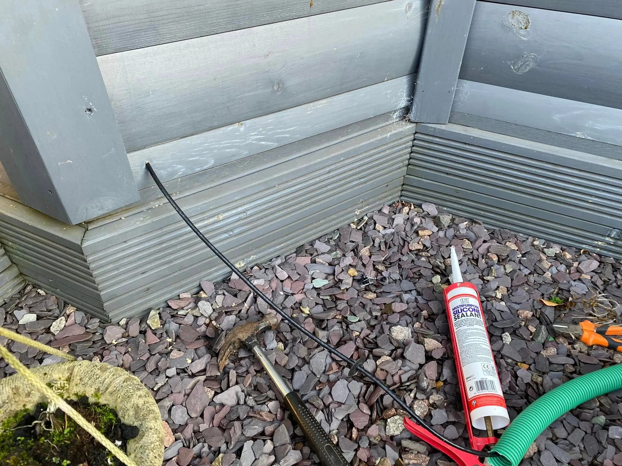 Black cable coming out of a grey timber cabin base onto slate chippings, with a claw hammer, silicone sealant and green conduit beside it