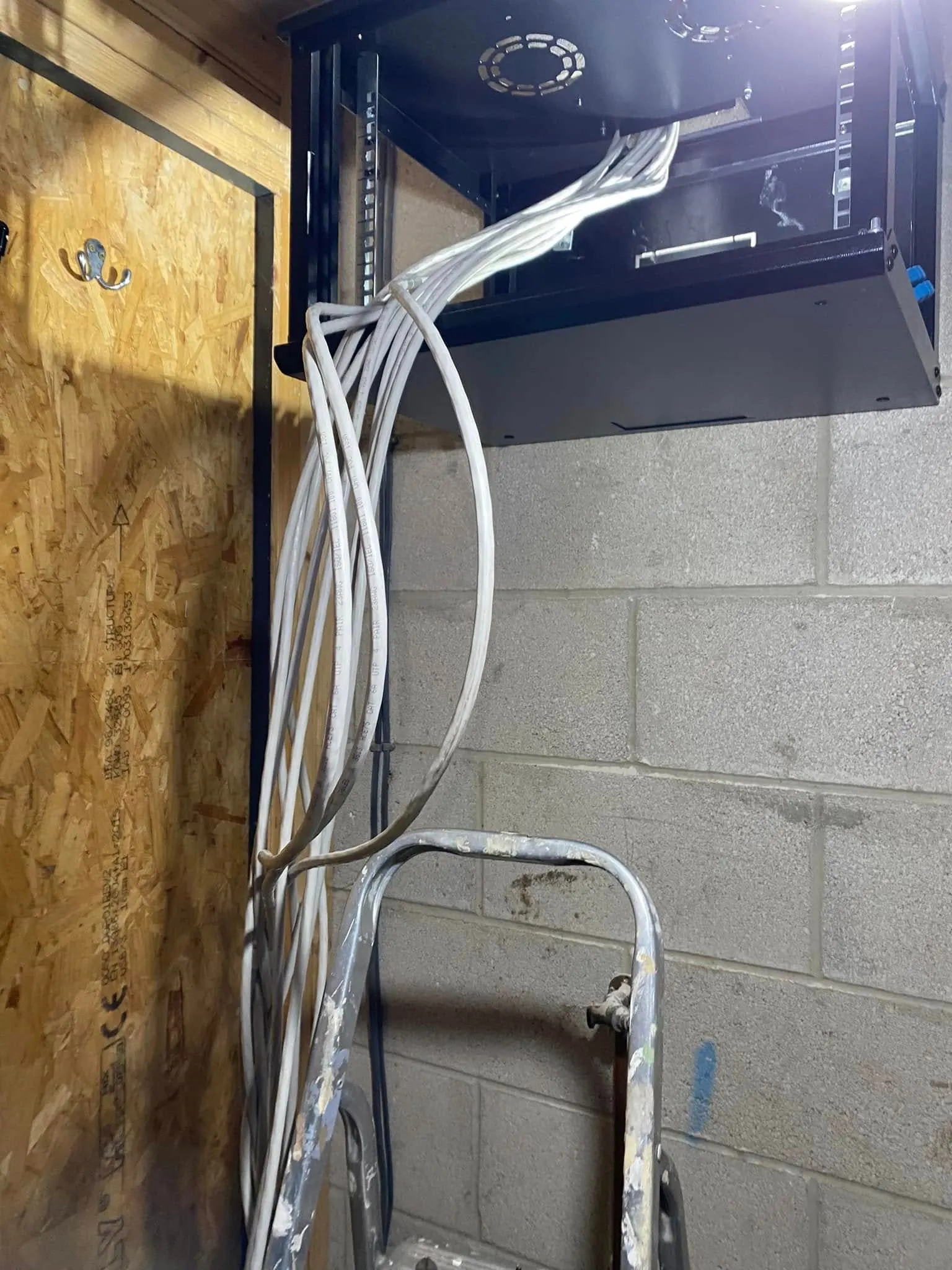 Bundle of loose white CAT6 cables hangs from a black wall-mounted network cabinet down a blockwork wall past a stepladder