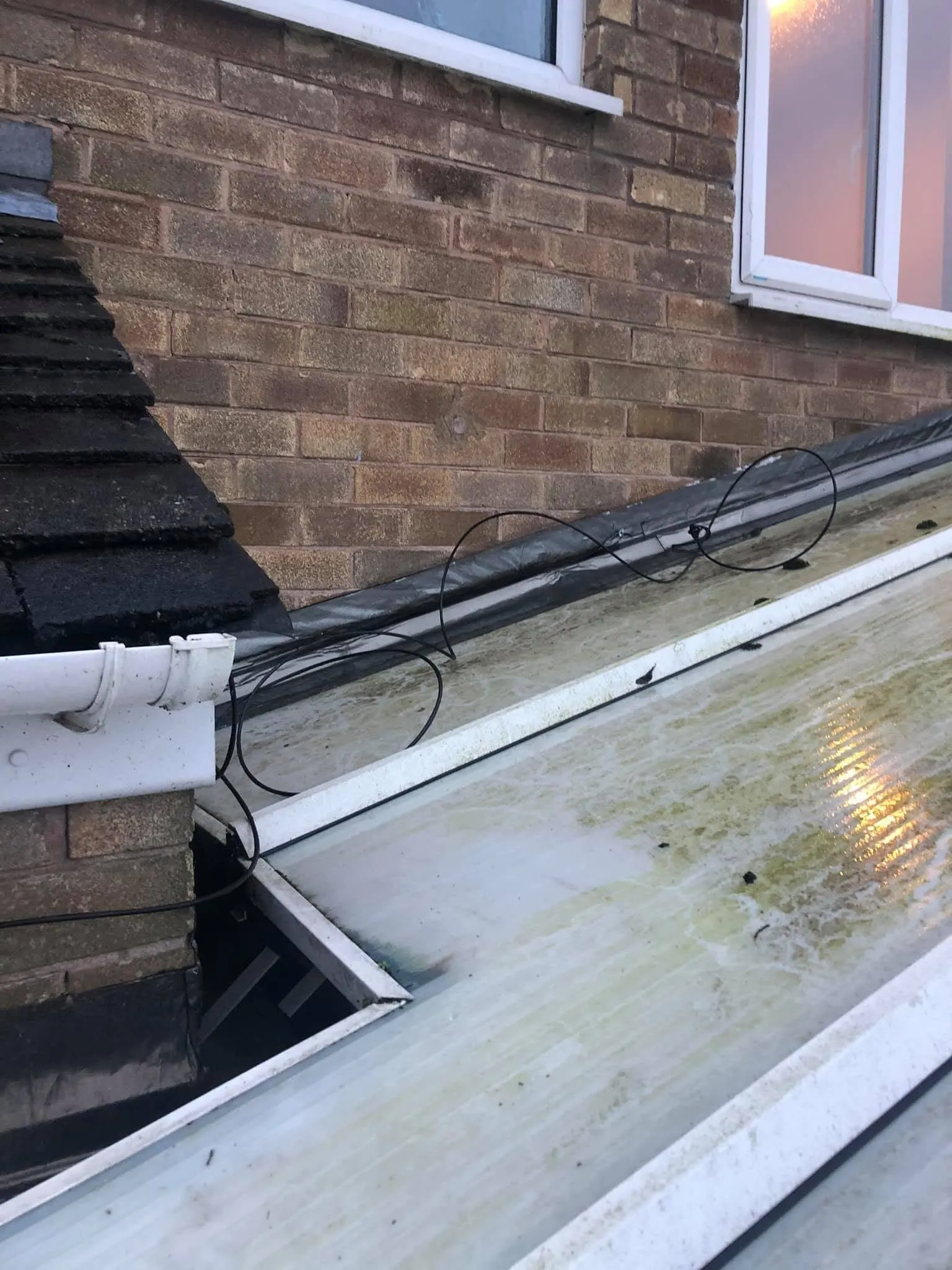 Black cable looped loosely across a conservatory roof below a brick wall, with a white gutter and roof tiles to the left