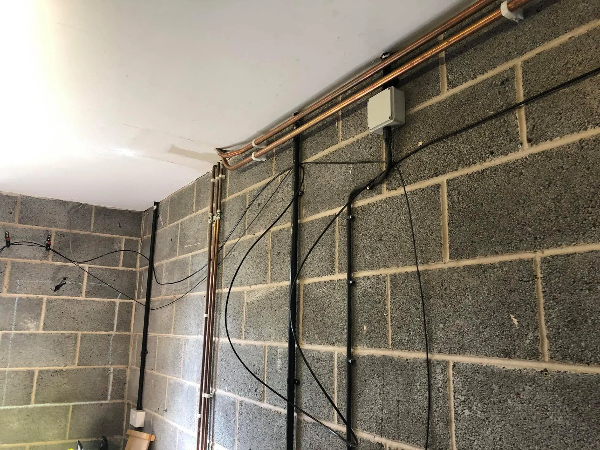 Black cables drape loosely across a grey blockwork garage wall below copper pipes and a grey junction box