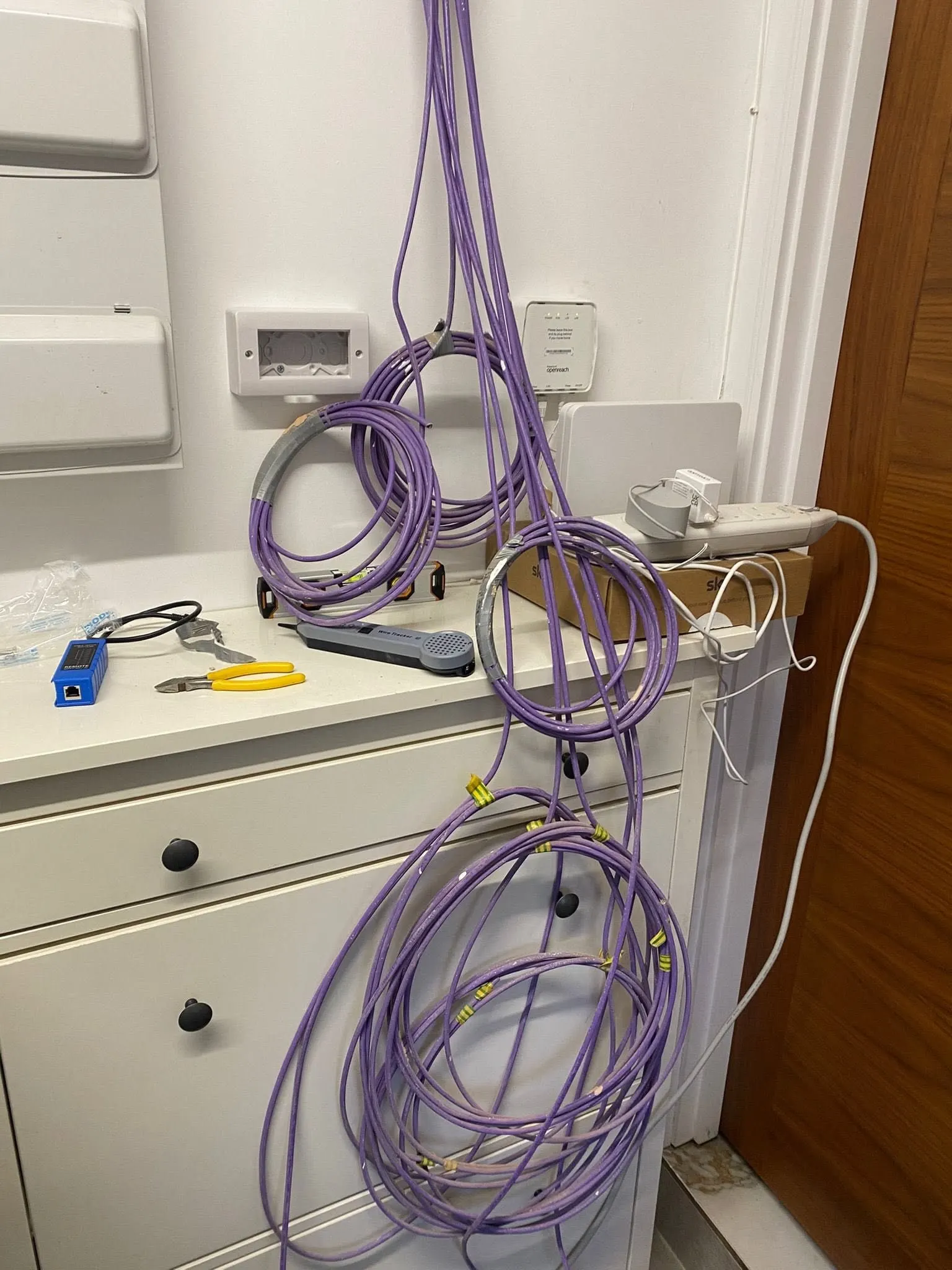Coils of purple network cable hang beside a consumer unit and an Openreach box above a chest of drawers with cable tools