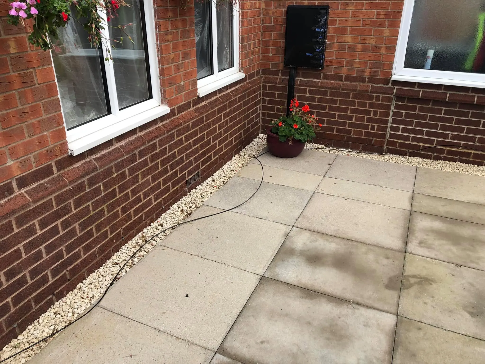 Loose black cable running across patio slabs and along a gravel strip beside a red brick wall, past a potted red geranium