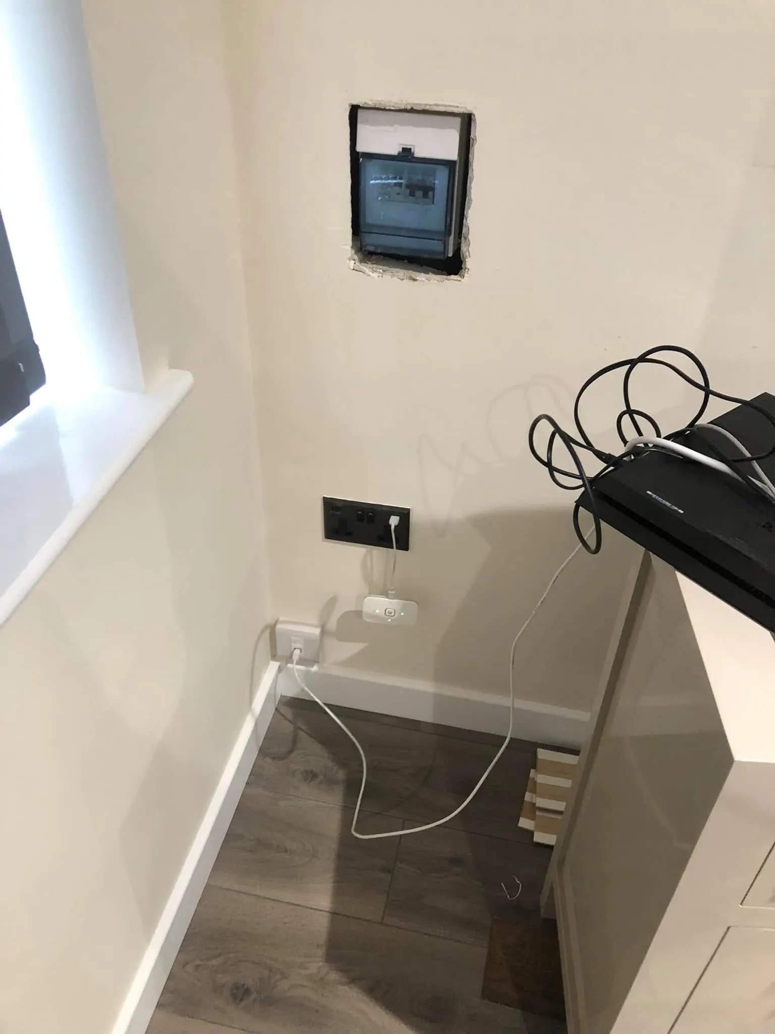 Black router balanced on a white cabinet with tangled black leads, near a plug socket, a phone socket and a fuse box set in the wall