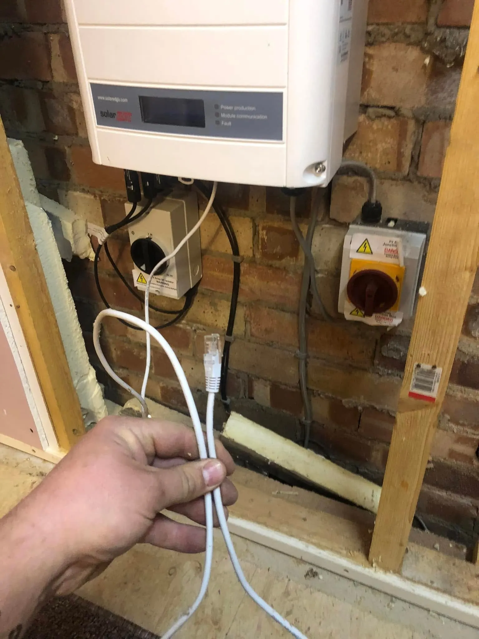Hand holding a white ethernet patch lead with an RJ45 plug below a SolarEdge solar inverter on a bare brick wall