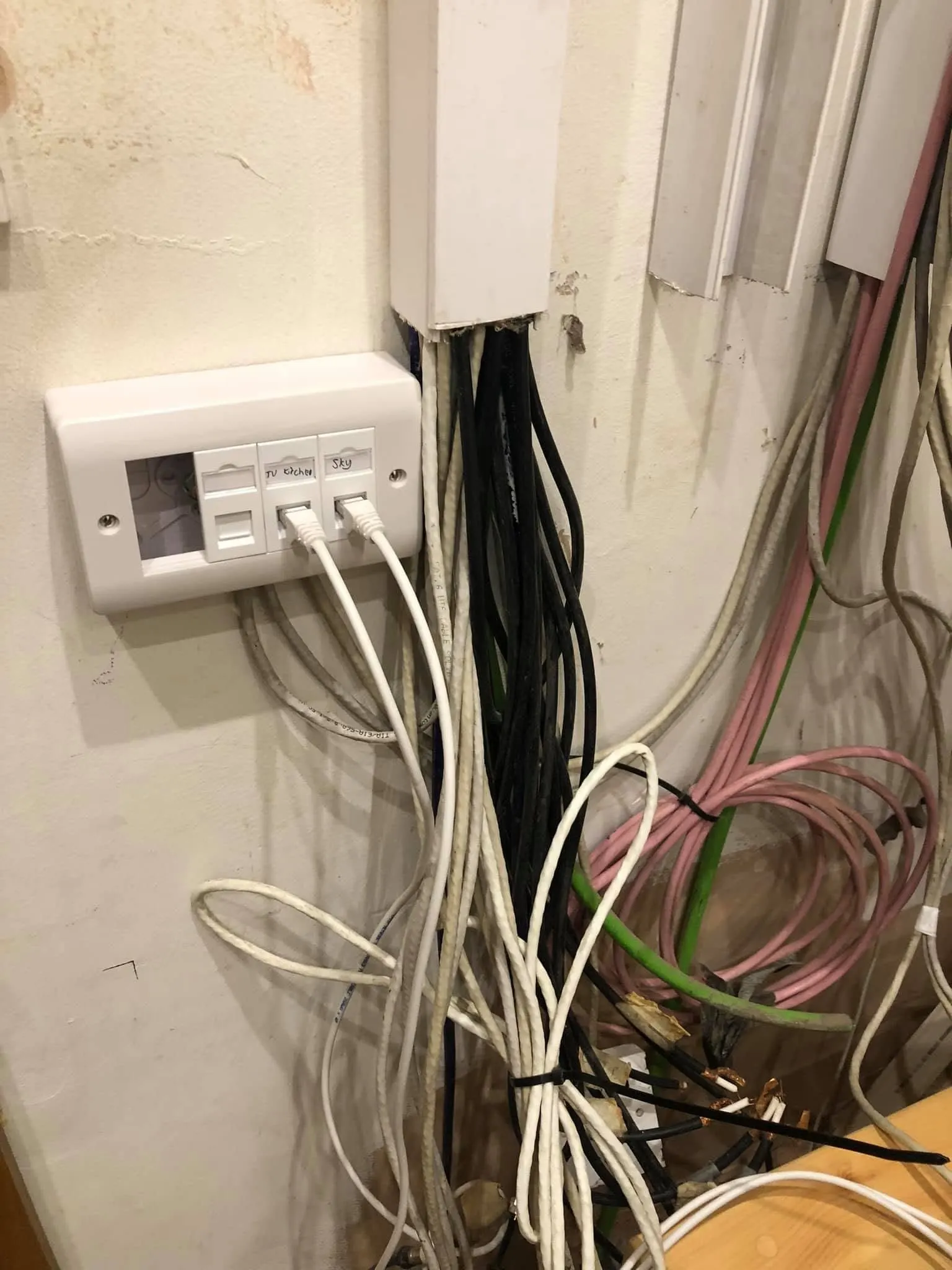 Mass of white, black and pink cables hanging from open trunking beside a faceplate hand-labelled TV Kitchen and Sky