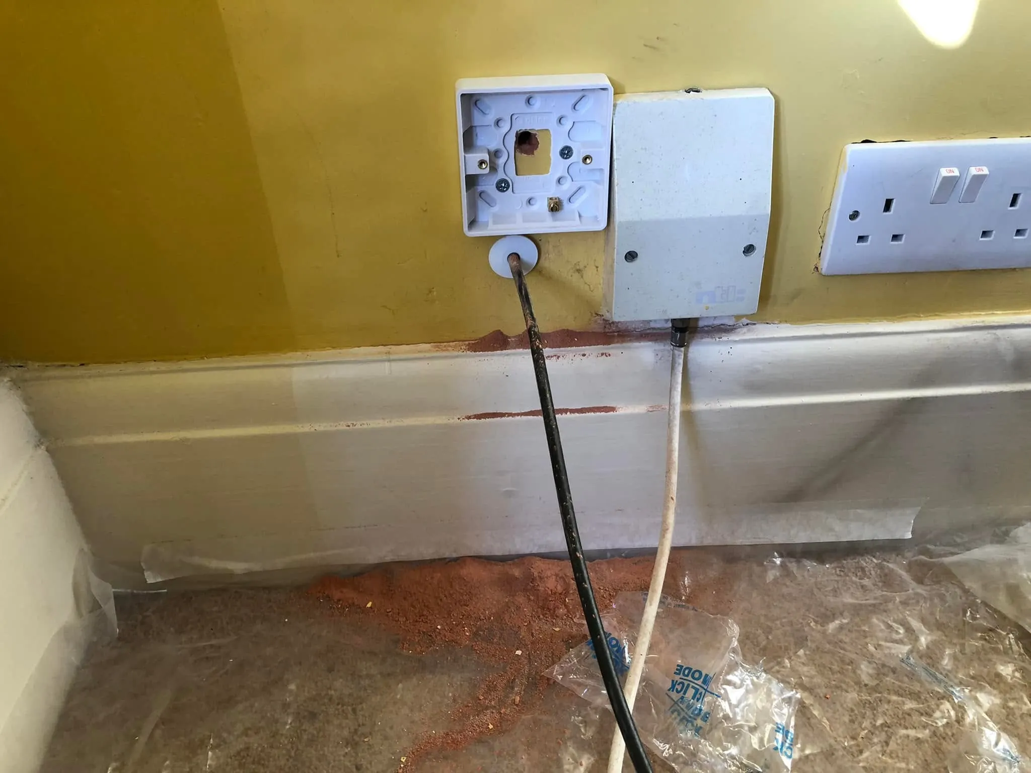 Empty white back box above a black cable in a grommet on a yellow wall, next to an NTL box, with brick dust on the skirting