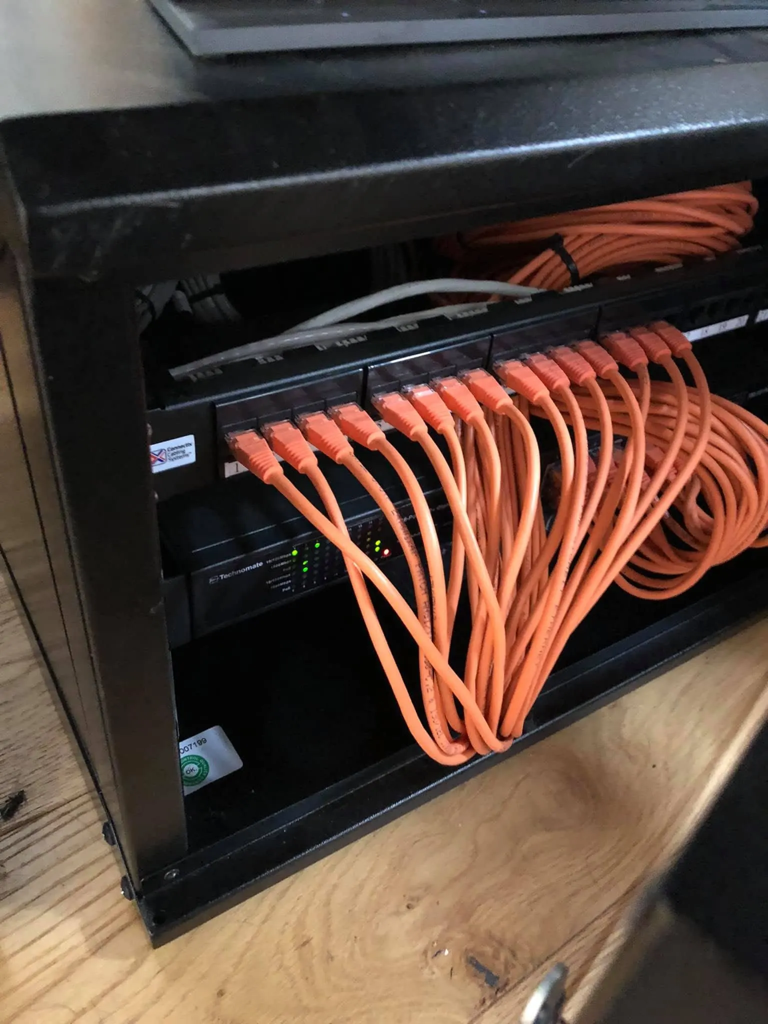 Orange patch leads looping from a patch panel down to a Technomate PoE switch inside a black wall cabinet