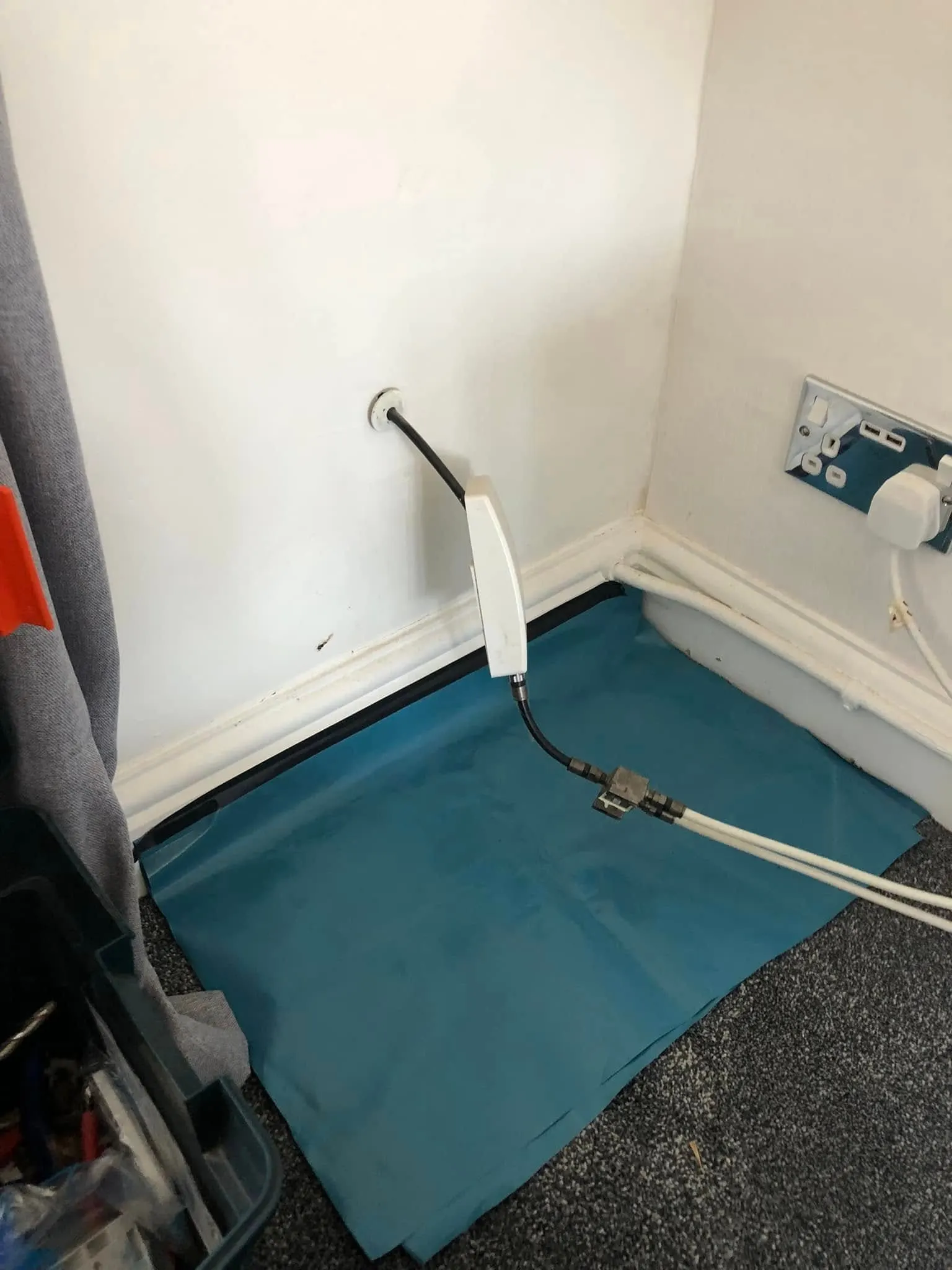Coax cable from a wall grommet runs through a white inline box to a metal splitter feeding two white leads on a blue dust sheet