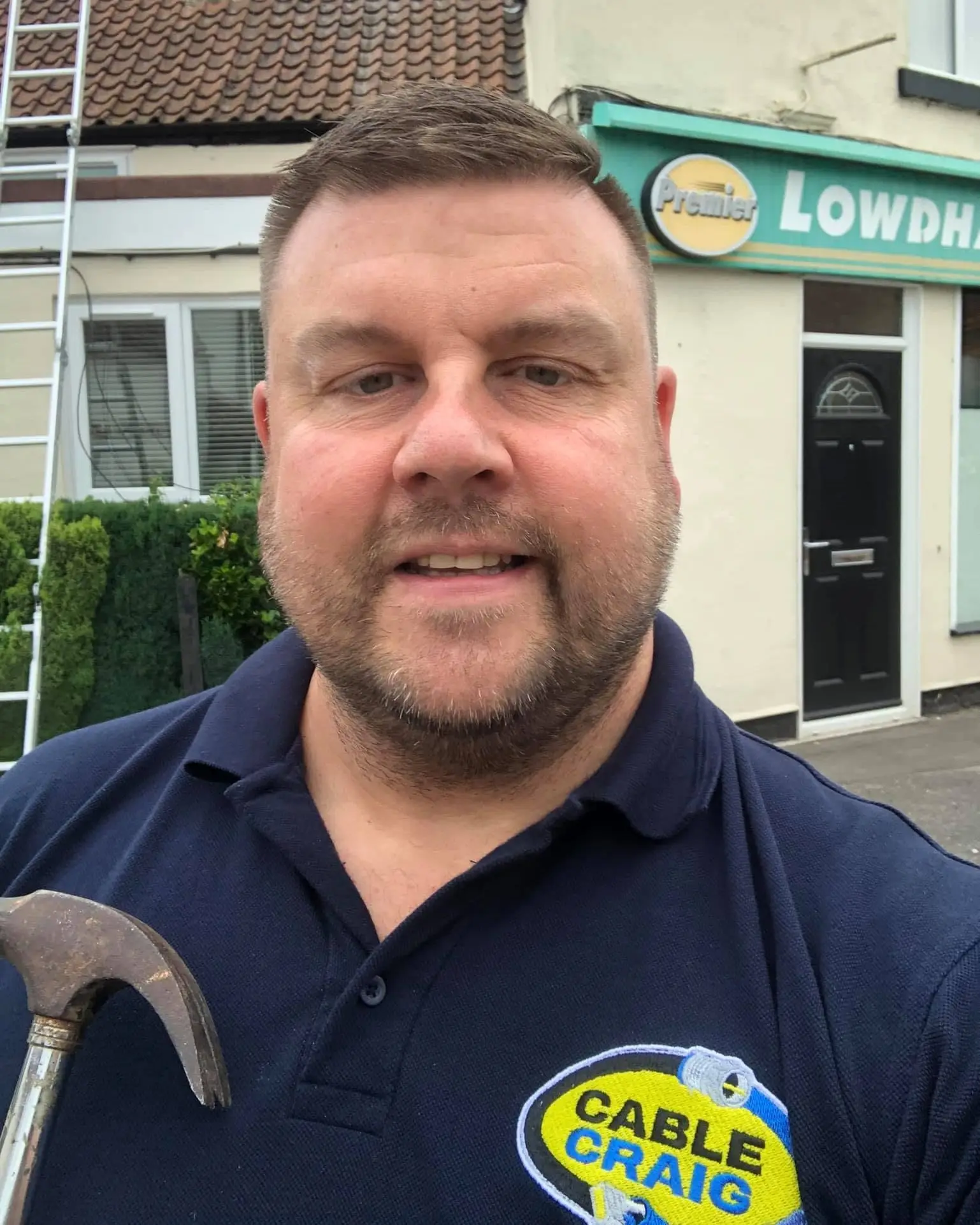 Craig in a navy Cable Craig polo holding a claw hammer outside a Premier shop in Lowdham, with a ladder against a cottage roof behind