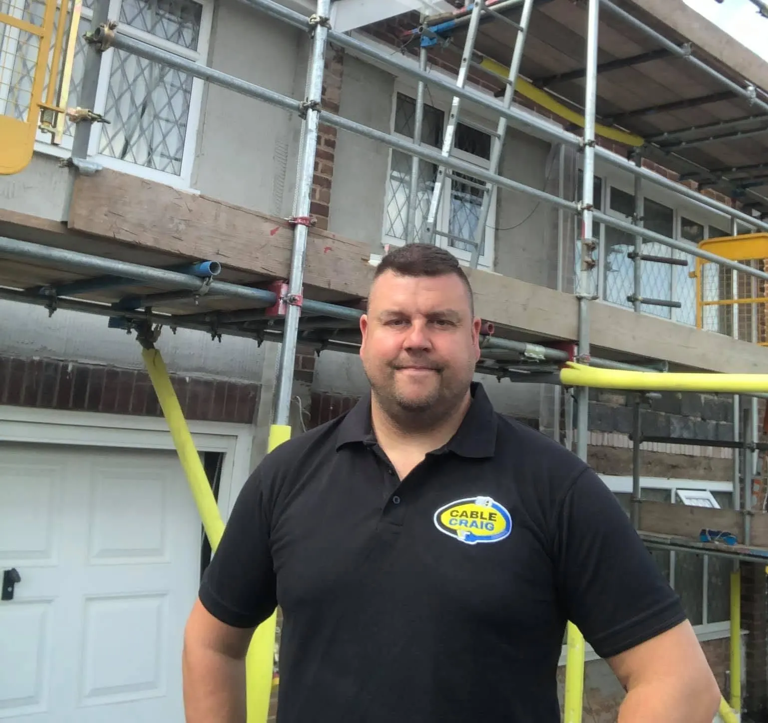 Craig in a black Cable Craig polo standing in front of a house wrapped in scaffolding with a ladder up to the roof