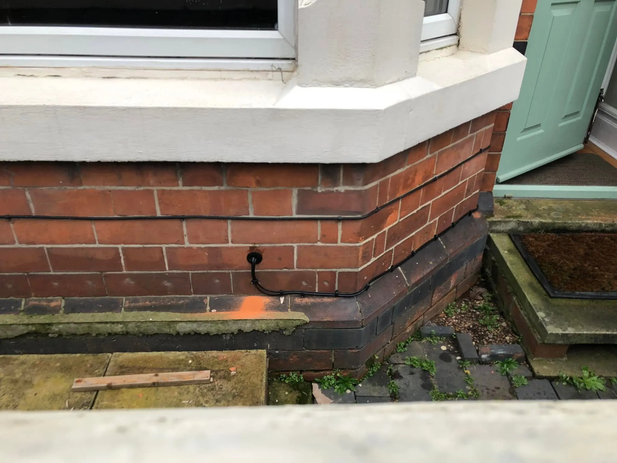 Black external cable clipped along the brick courses of a bay window and into a wall grommet beside a green front door
