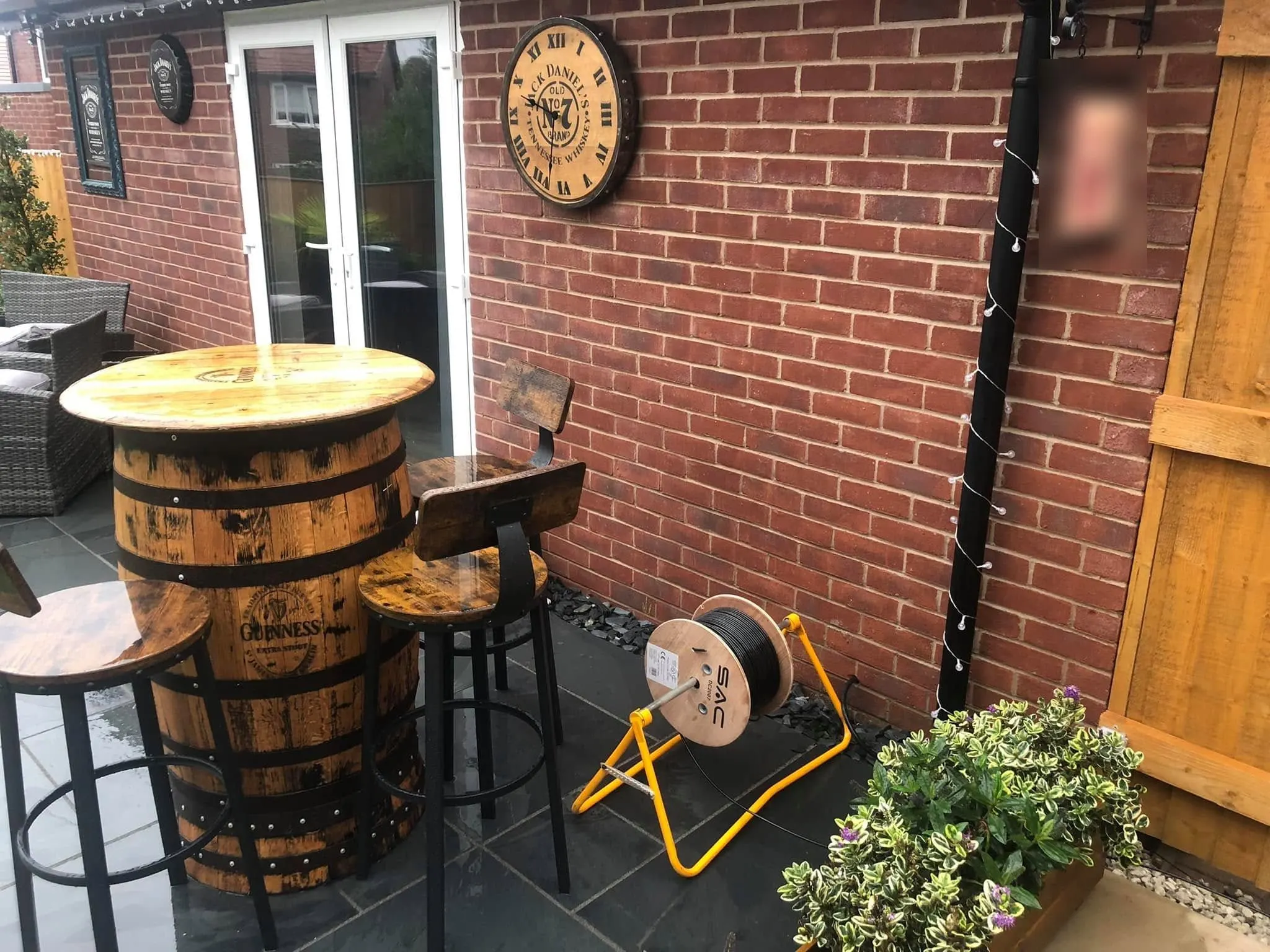 Drum of black cable on a yellow reel stand on a slate patio beside a barrel table, bar stools and a red brick wall
