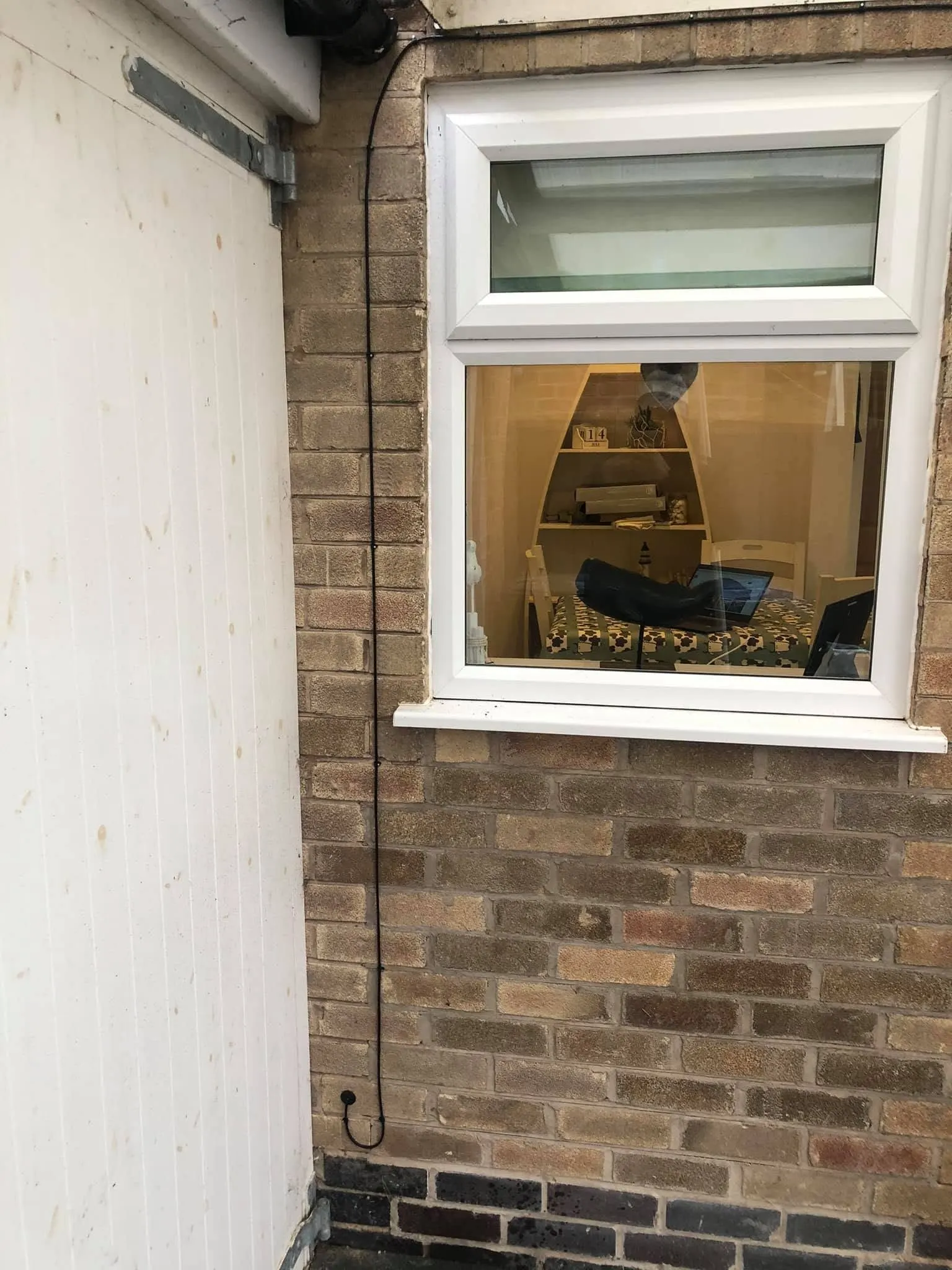 Thin black cable running down buff brick beside a white window, forming a drip loop into a grommet near the ground