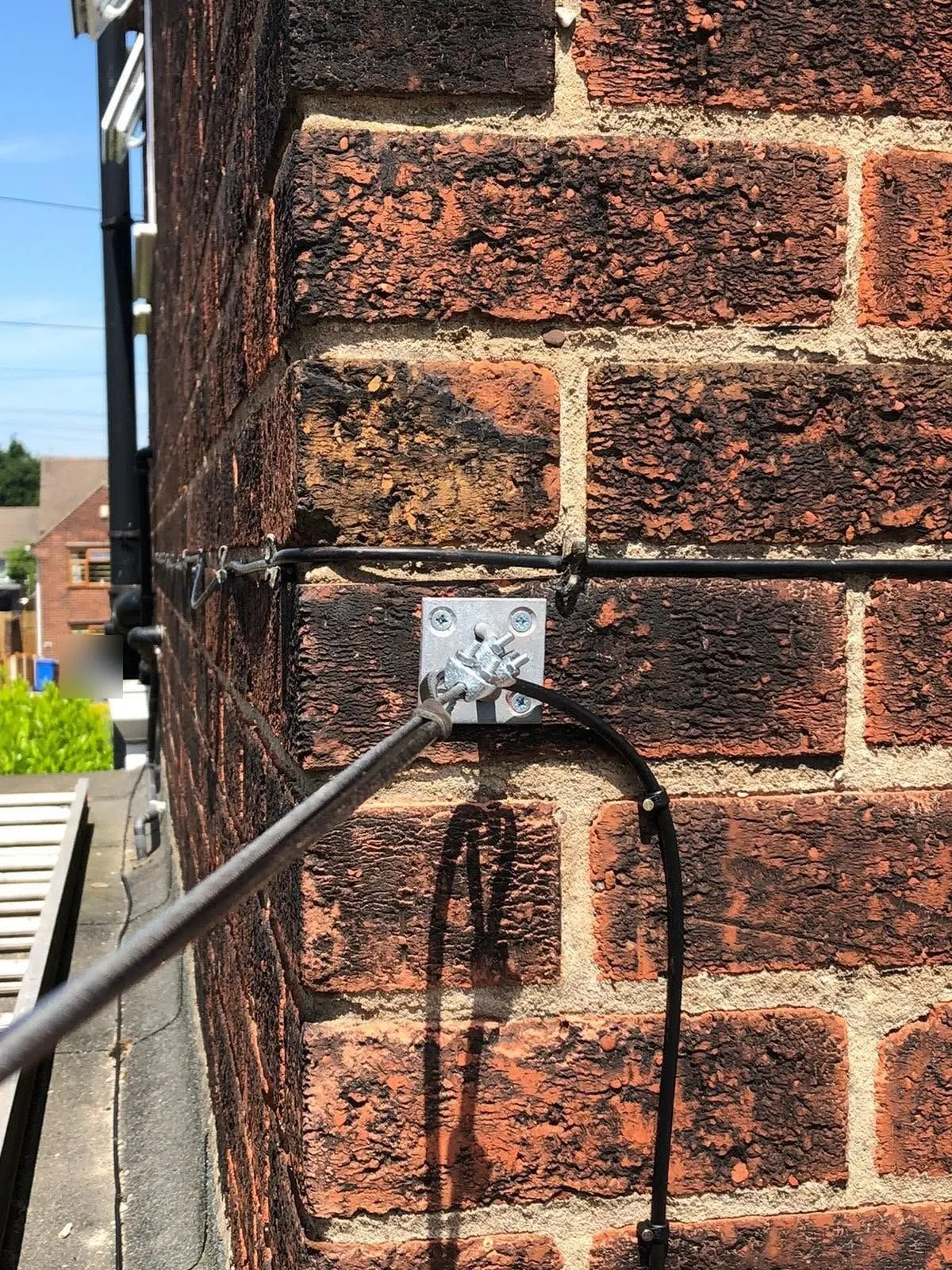 Galvanised anchor plate on a red brick corner holding a catenary wire, with a black cable clipped along the mortar line