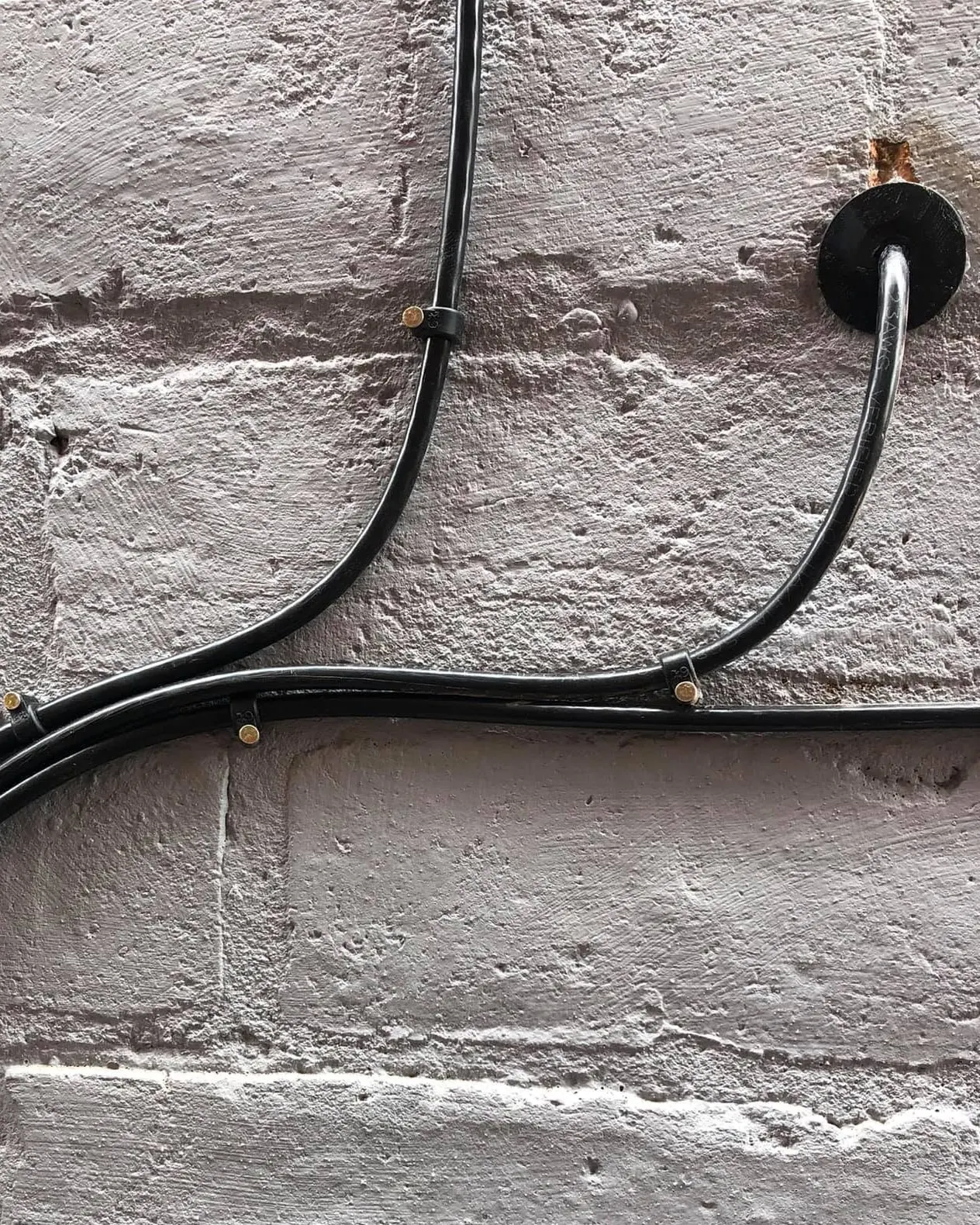 Black cables clipped in smooth sweeping curves across a grey painted brick wall, one feeding in through a rubber grommet
