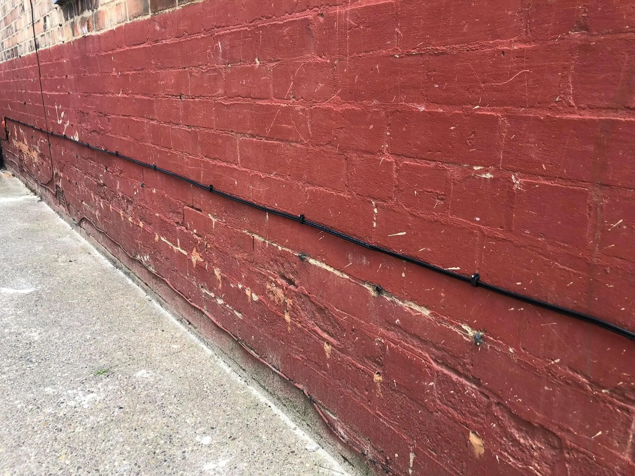 Black cable clipped in a long straight run along a painted red brick wall above a concrete path
