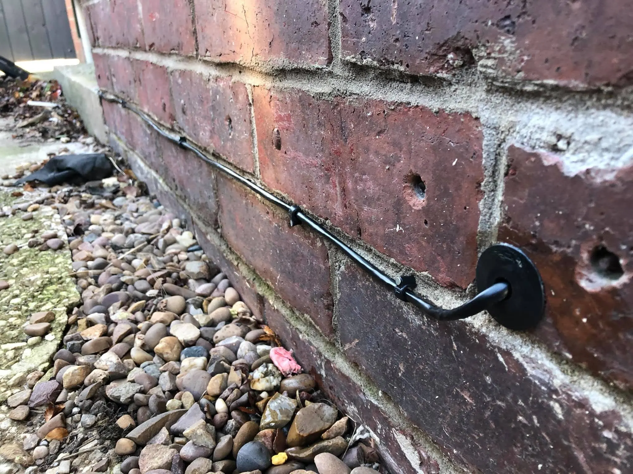 Black cable clipped along a mortar joint at the base of a brick wall, entering through a round black grommet above gravel