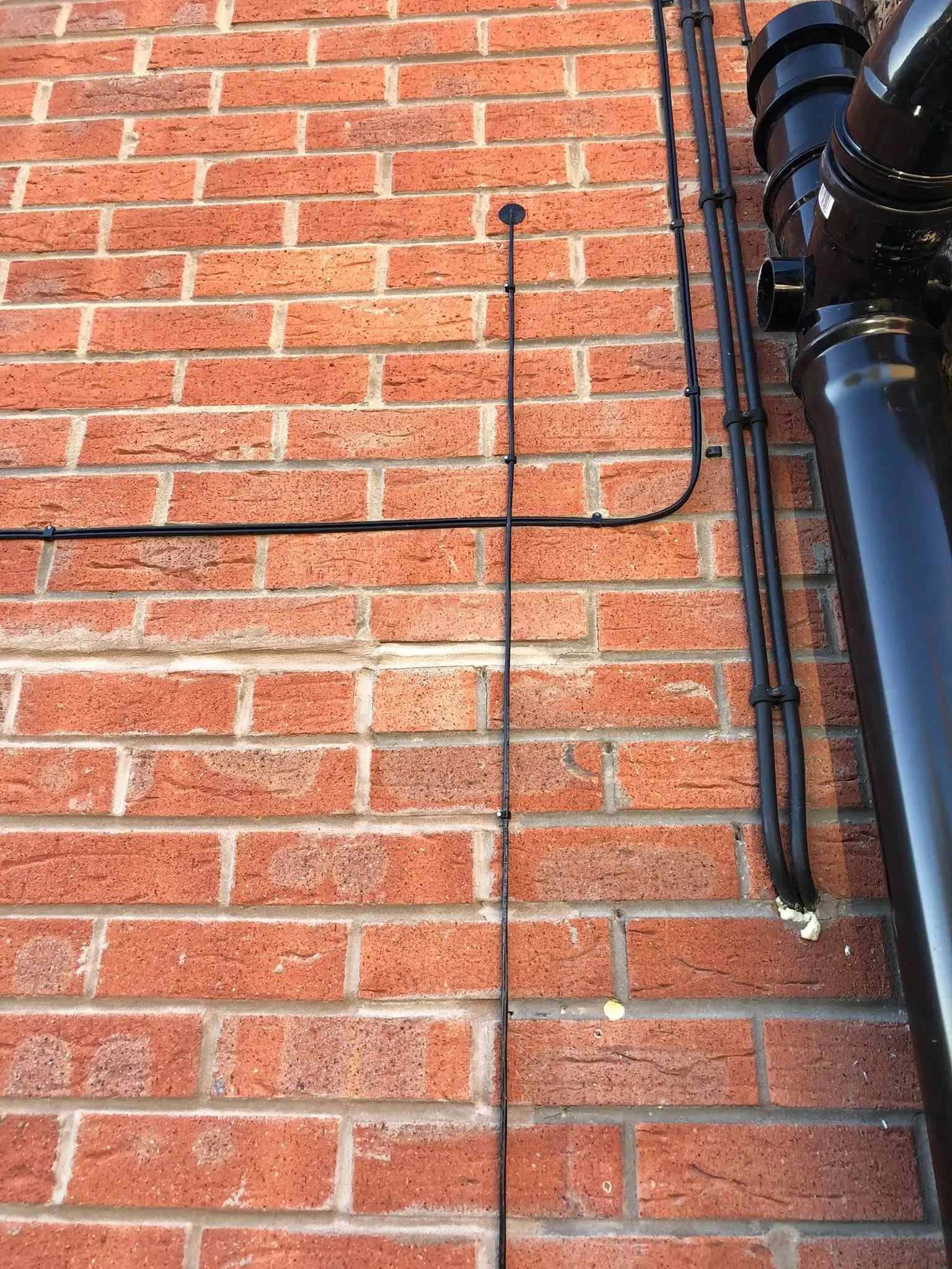 Black cables clipped in lines on orange brick beside a black downpipe, one ending at a round grommet