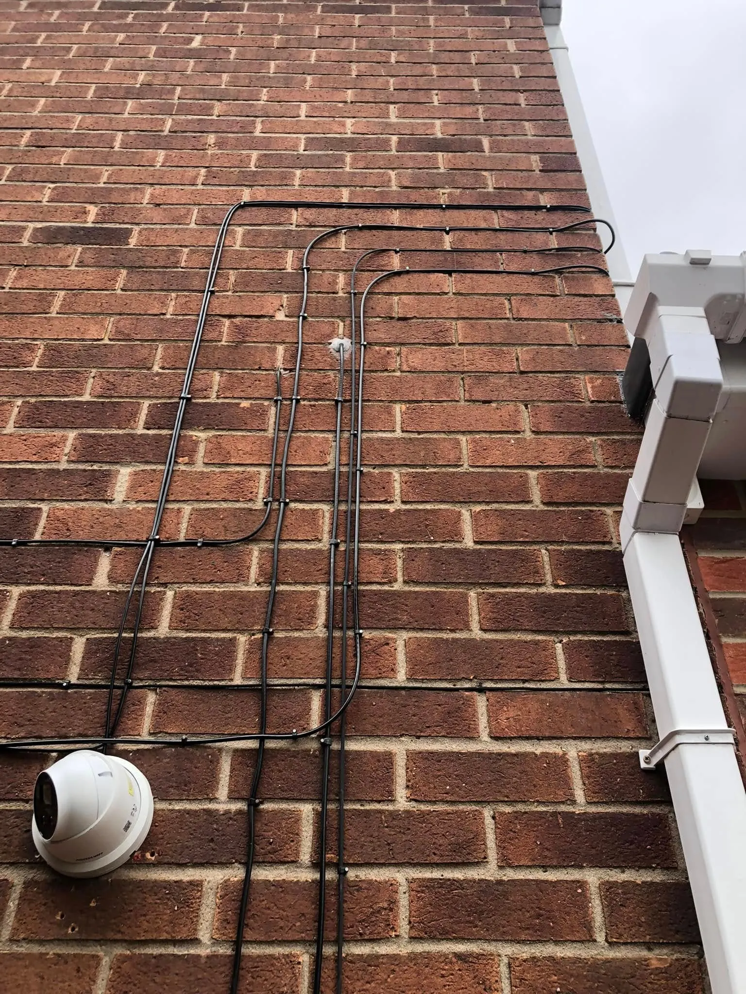 Several black cables running in parallel with rounded bends across red brick, feeding down past a white dome CCTV camera