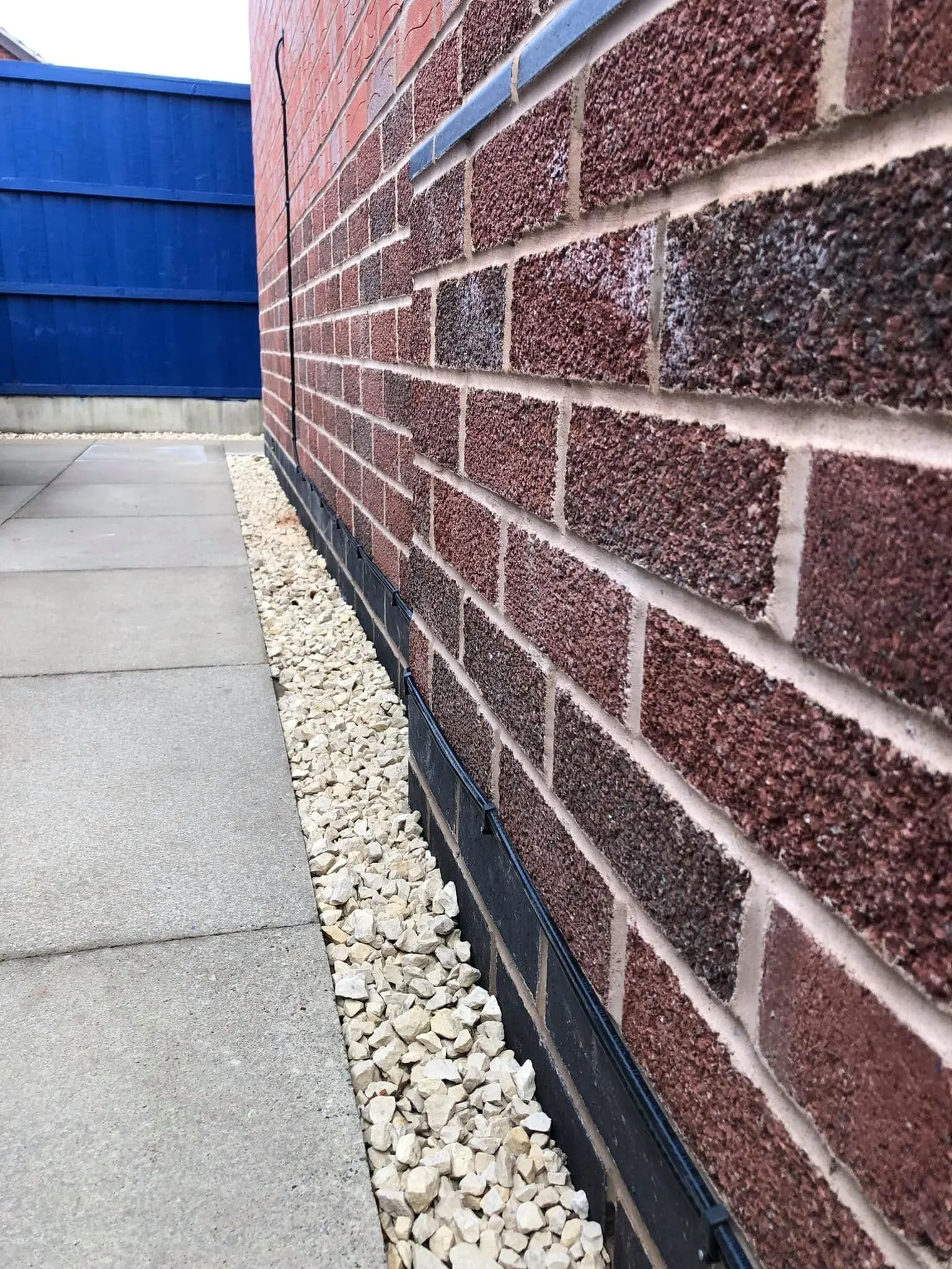 Black cable clipped along the dark plinth of a red brick wall above a strip of pale gravel, with a vertical drop further along