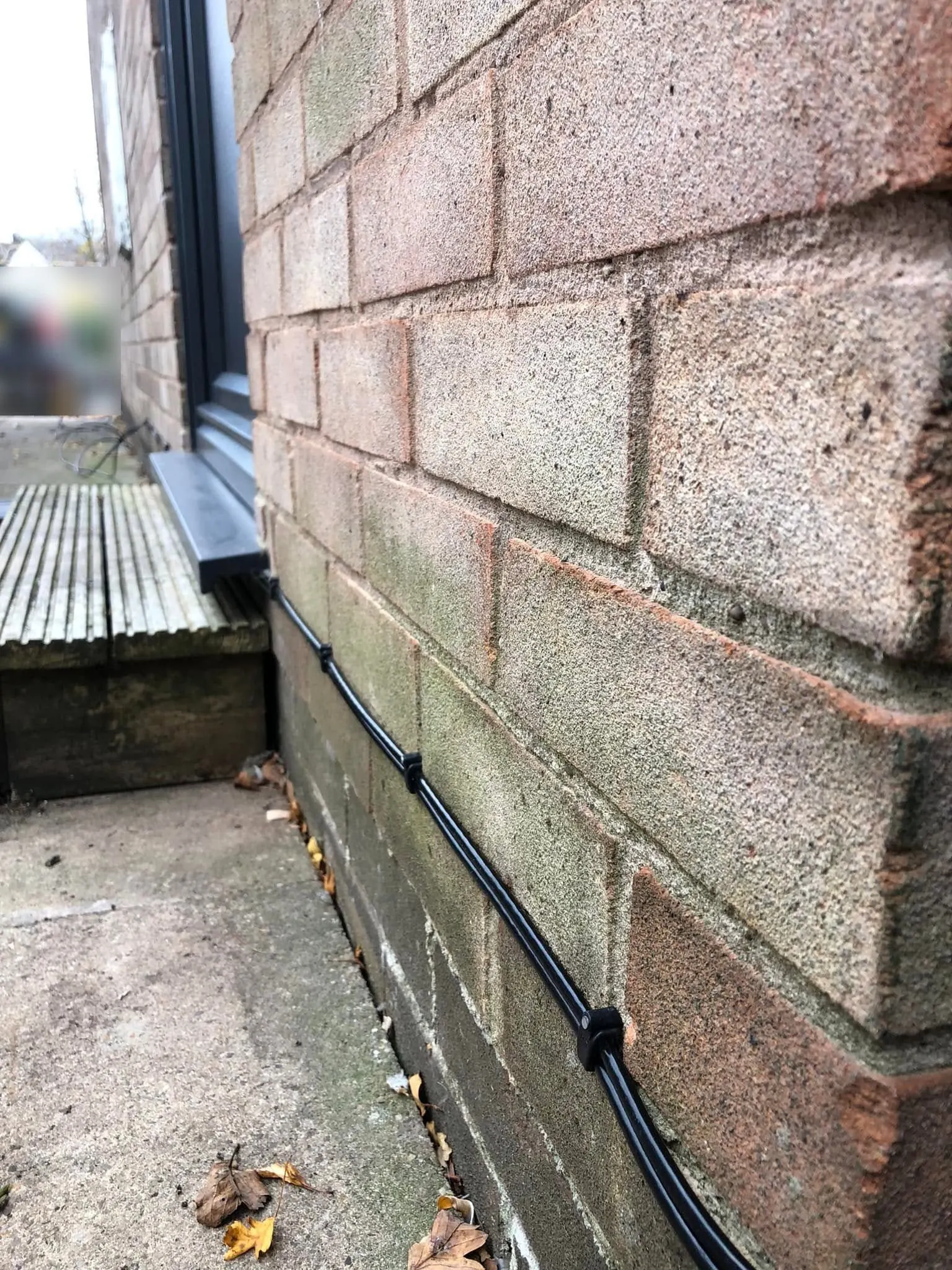 Black cable clipped along the plinth of a buff brick wall at ground level, running past timber decking