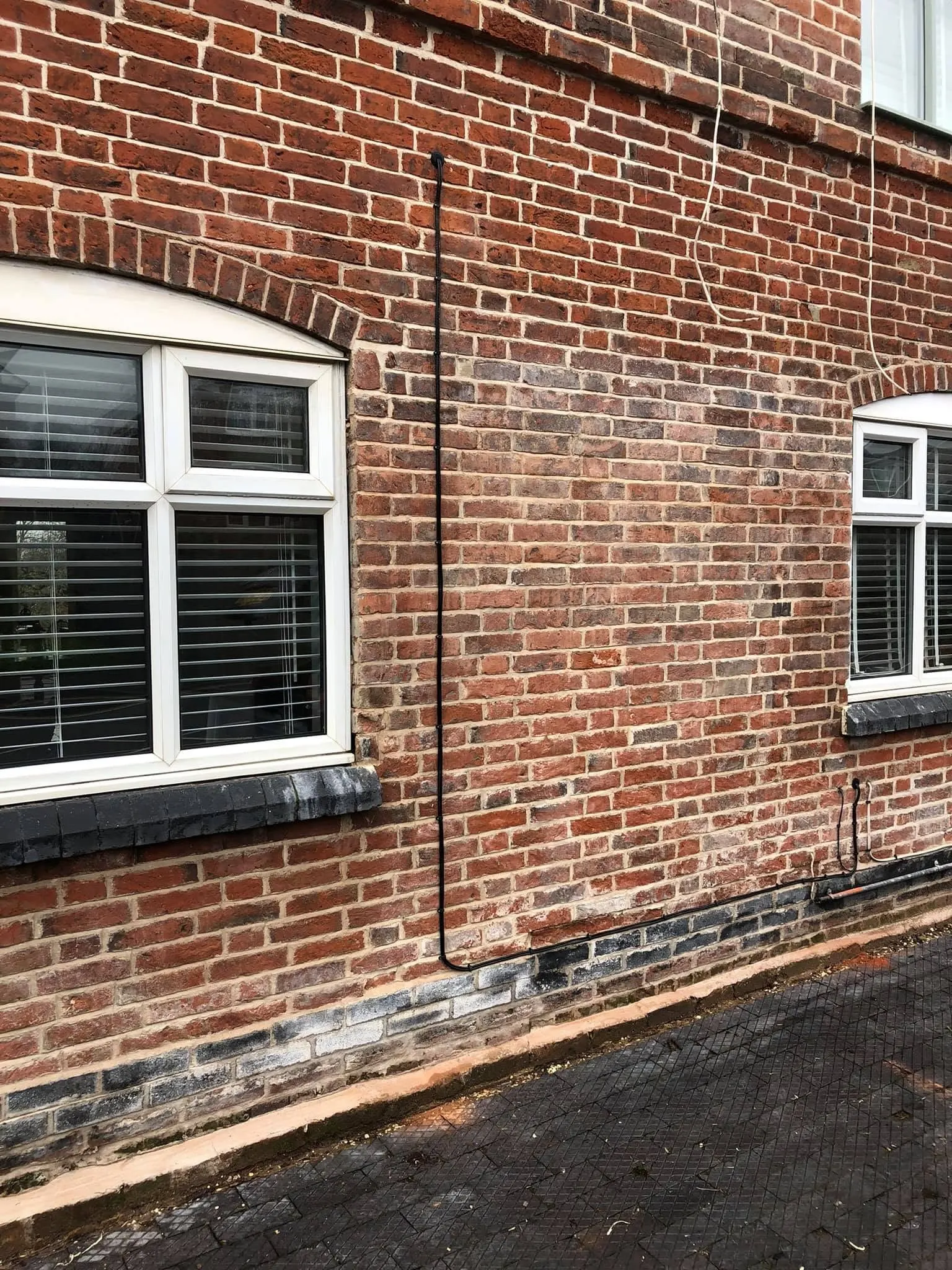 Black cable dropping from a grommet down a red brick facade between two windows, then running along a blue brick course