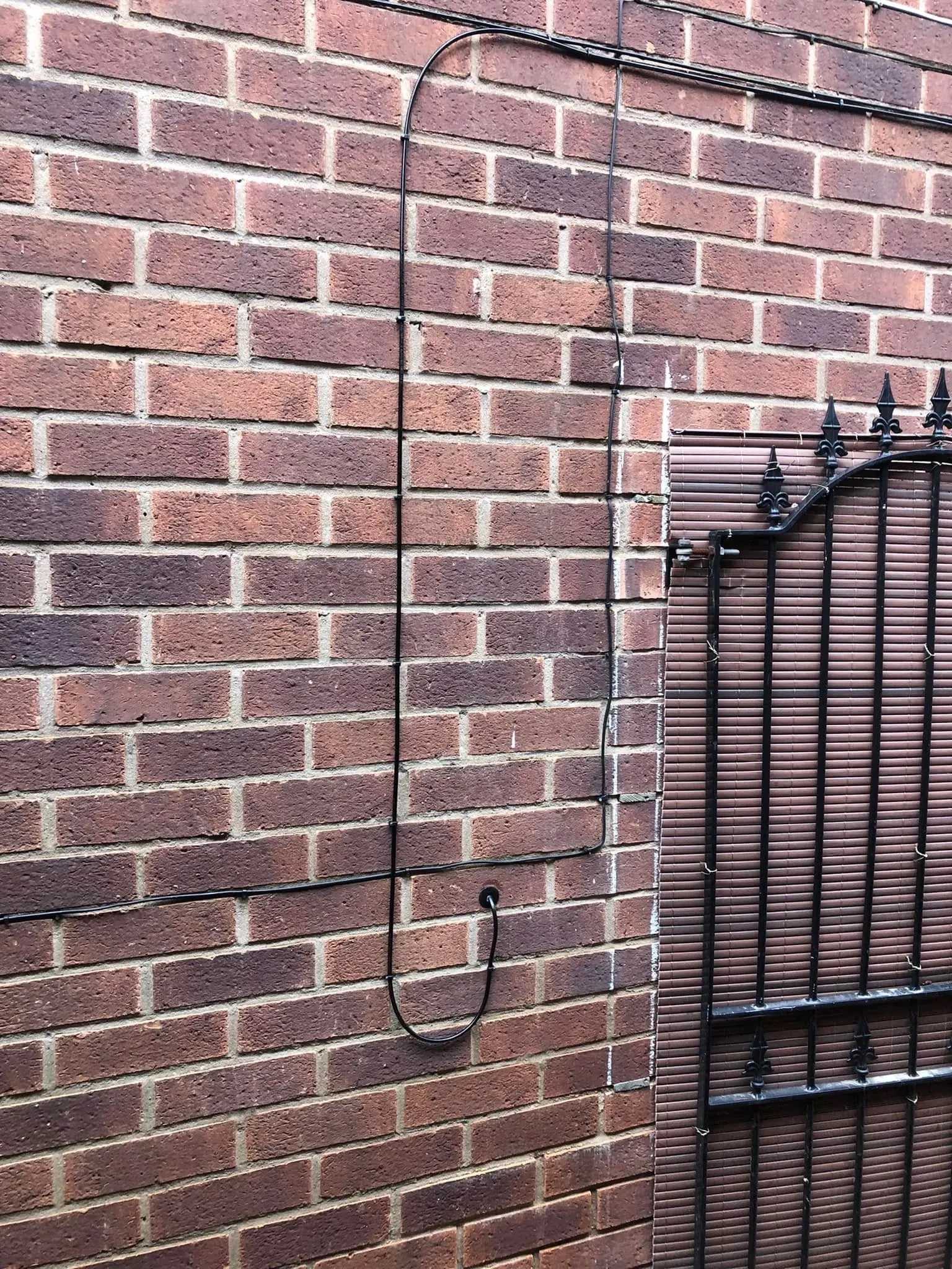 Black cables clipped with right-angle bends on brown brick, one dropping into a drip loop before a wall grommet, beside a metal gate