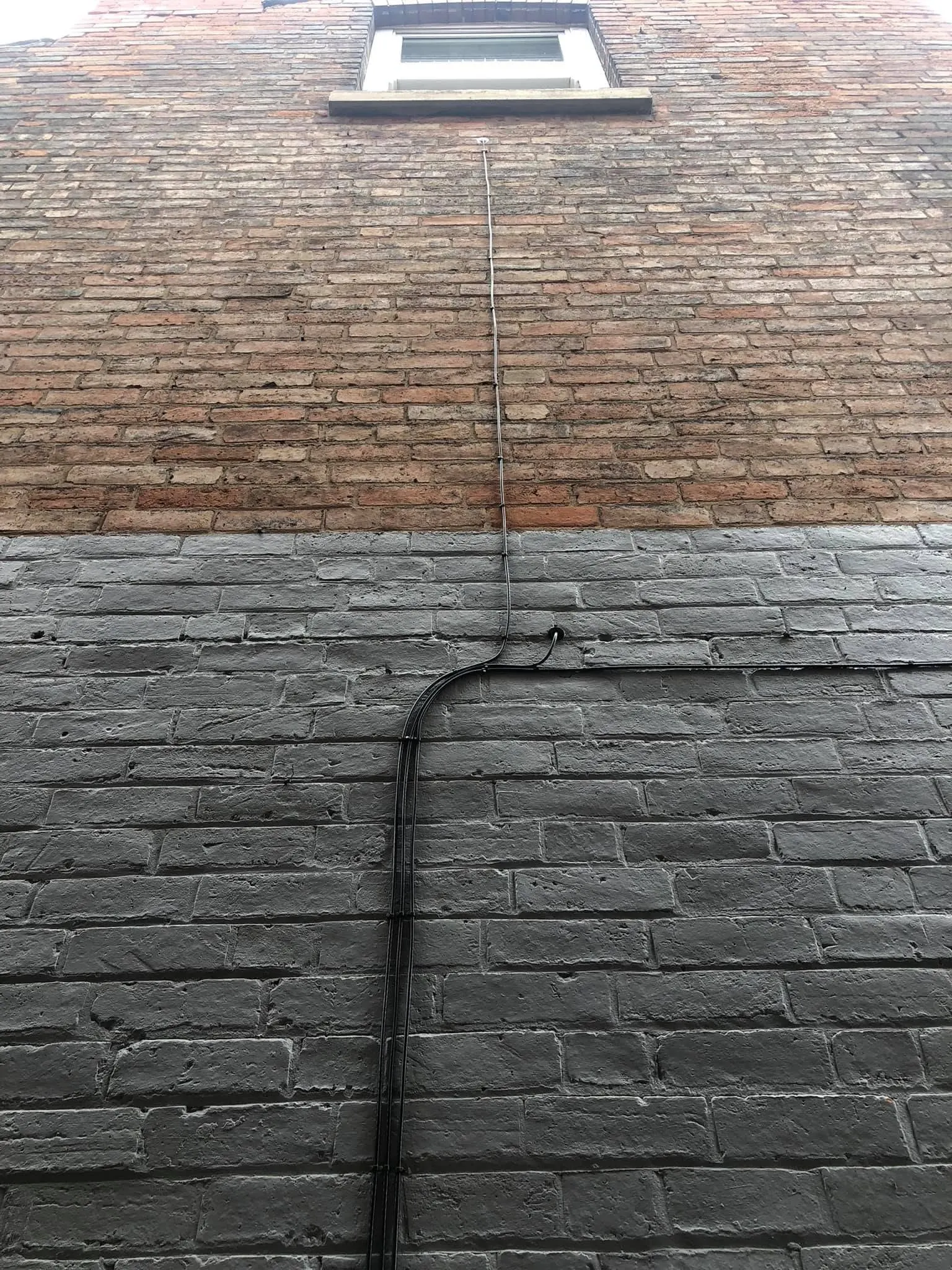 Tall brick wall with grey-painted lower courses, cables curving into a grommet and a thin cable rising to a window