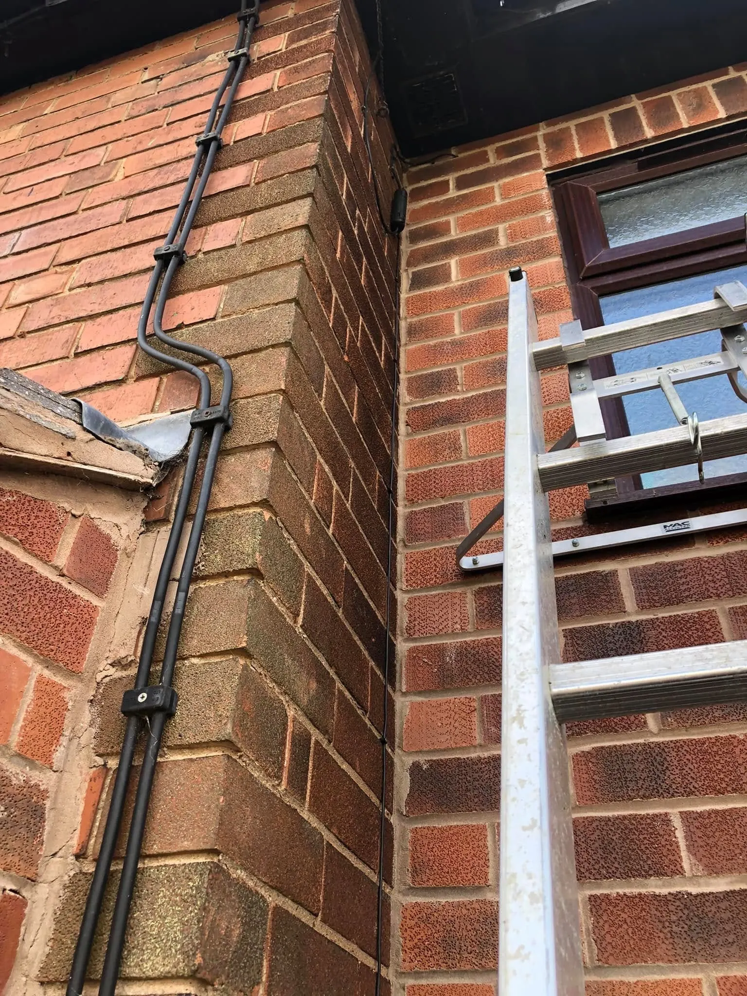 Two black cables clipped together down red brick, jogging round lead flashing, with a ladder and stand-off against the wall