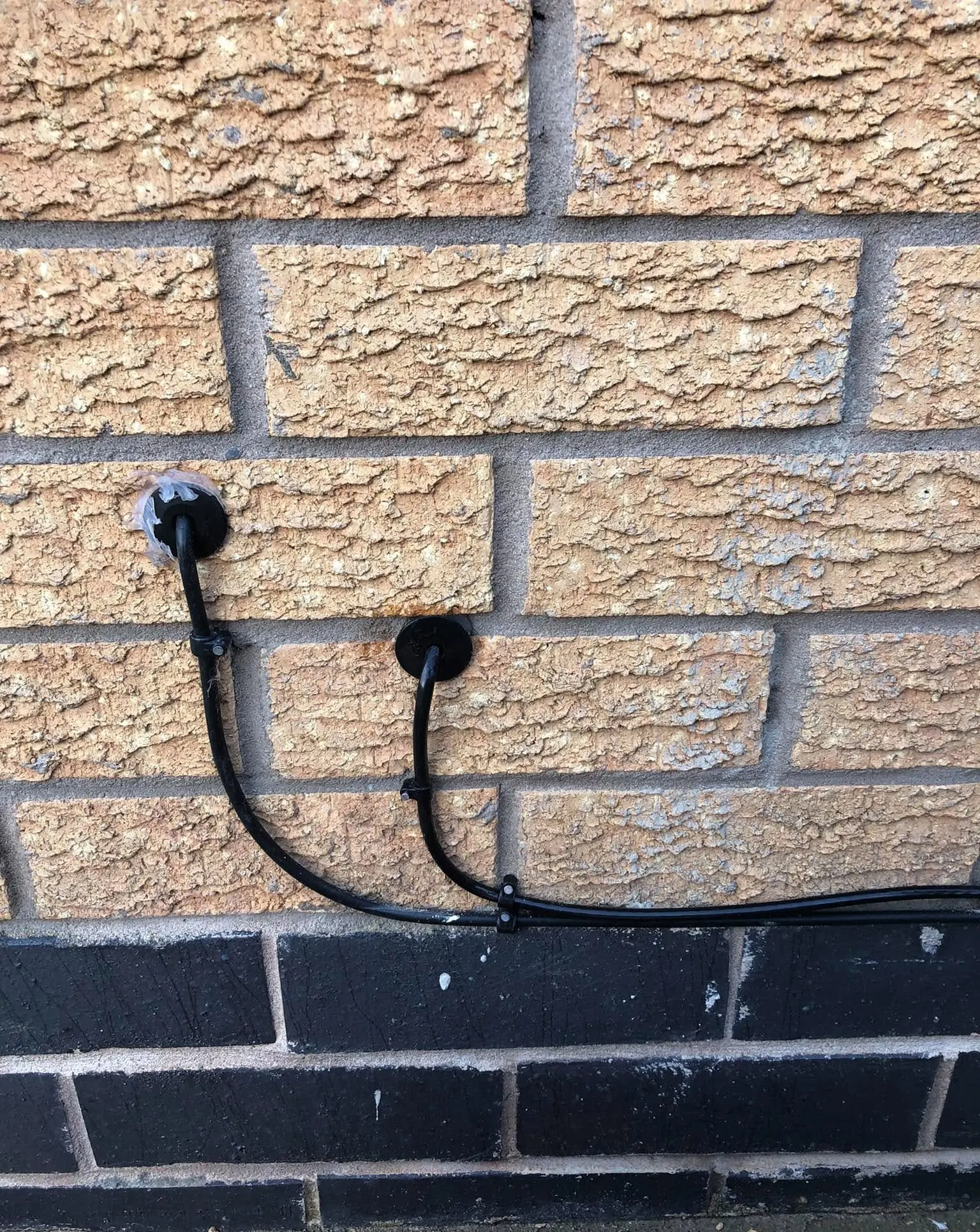 Two black cables entering buff brick through round grommets, curving down to join a run along a black brick course