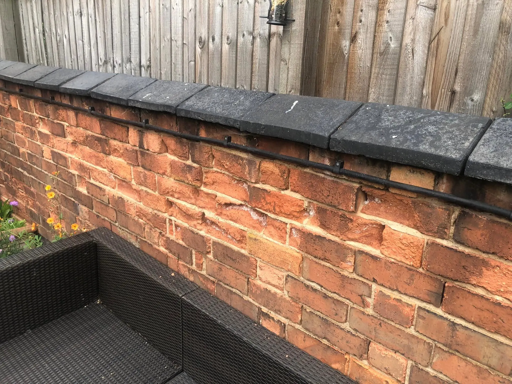 Black cable clipped along an old red brick garden wall just under grey coping stones, above rattan garden furniture