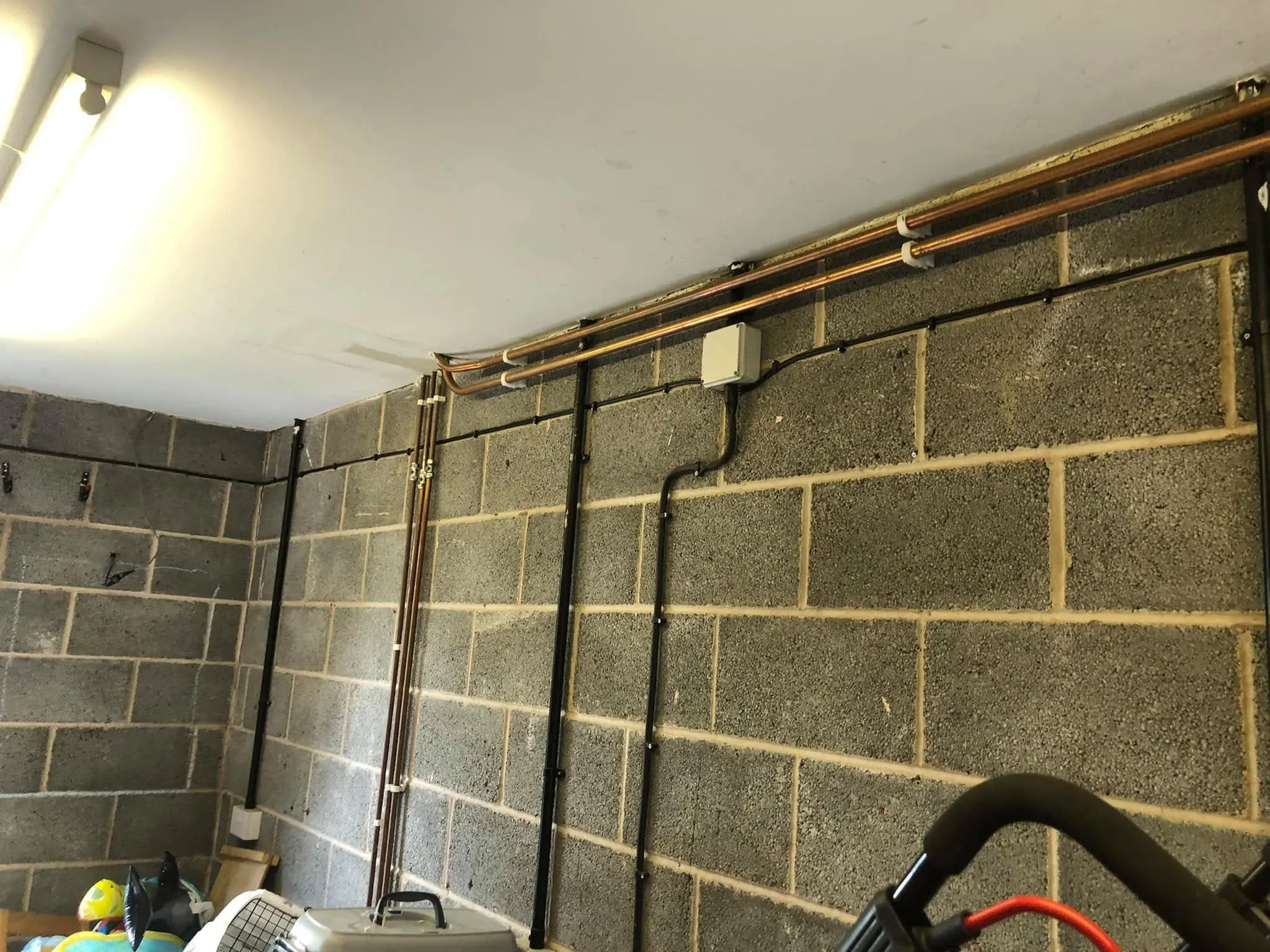 Black cables clipped in level runs across a grey blockwork garage wall beneath copper pipes and a grey junction box