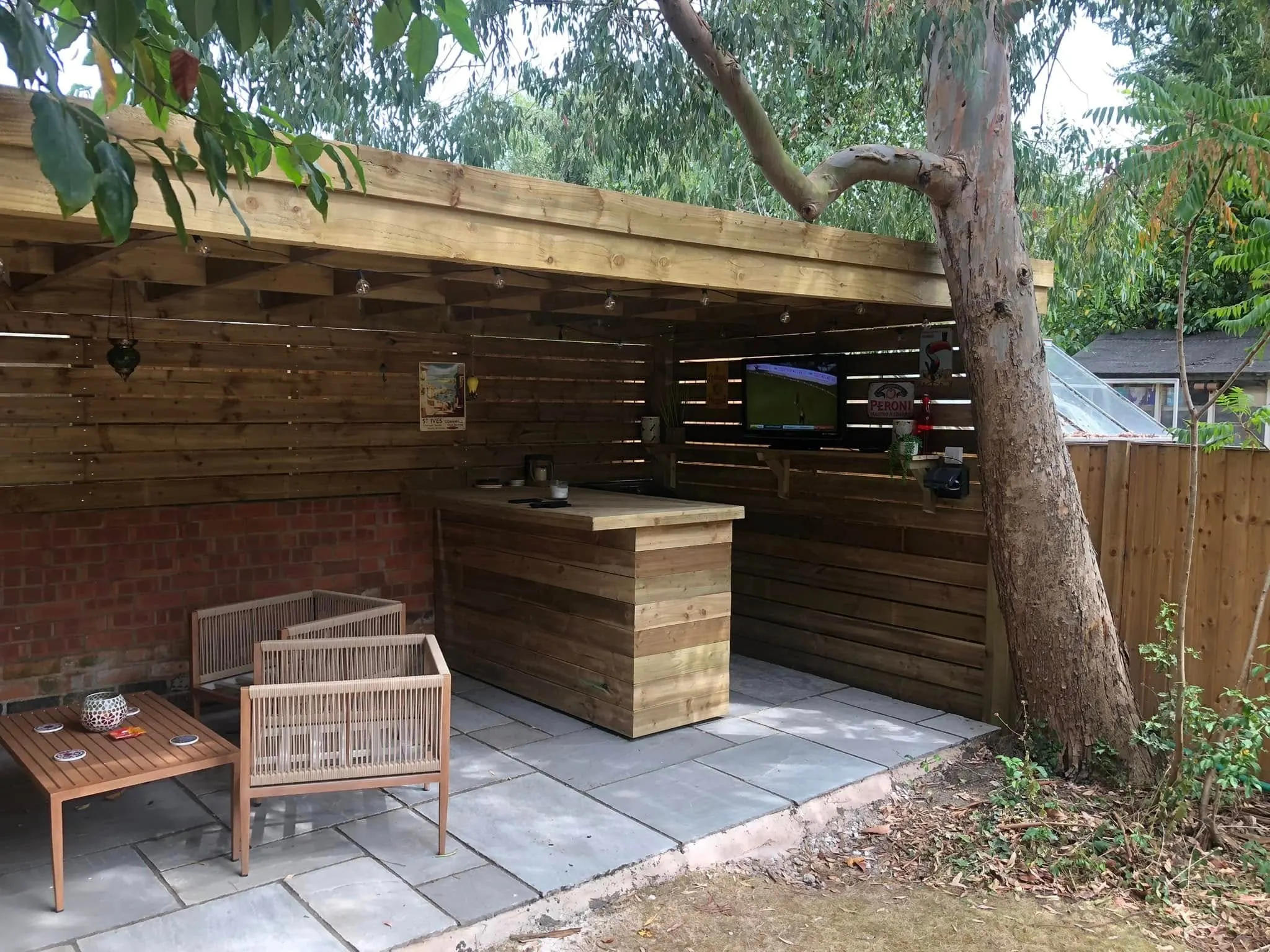 Covered timber garden bar with rope armchairs, a counter and a TV on the back wall showing football