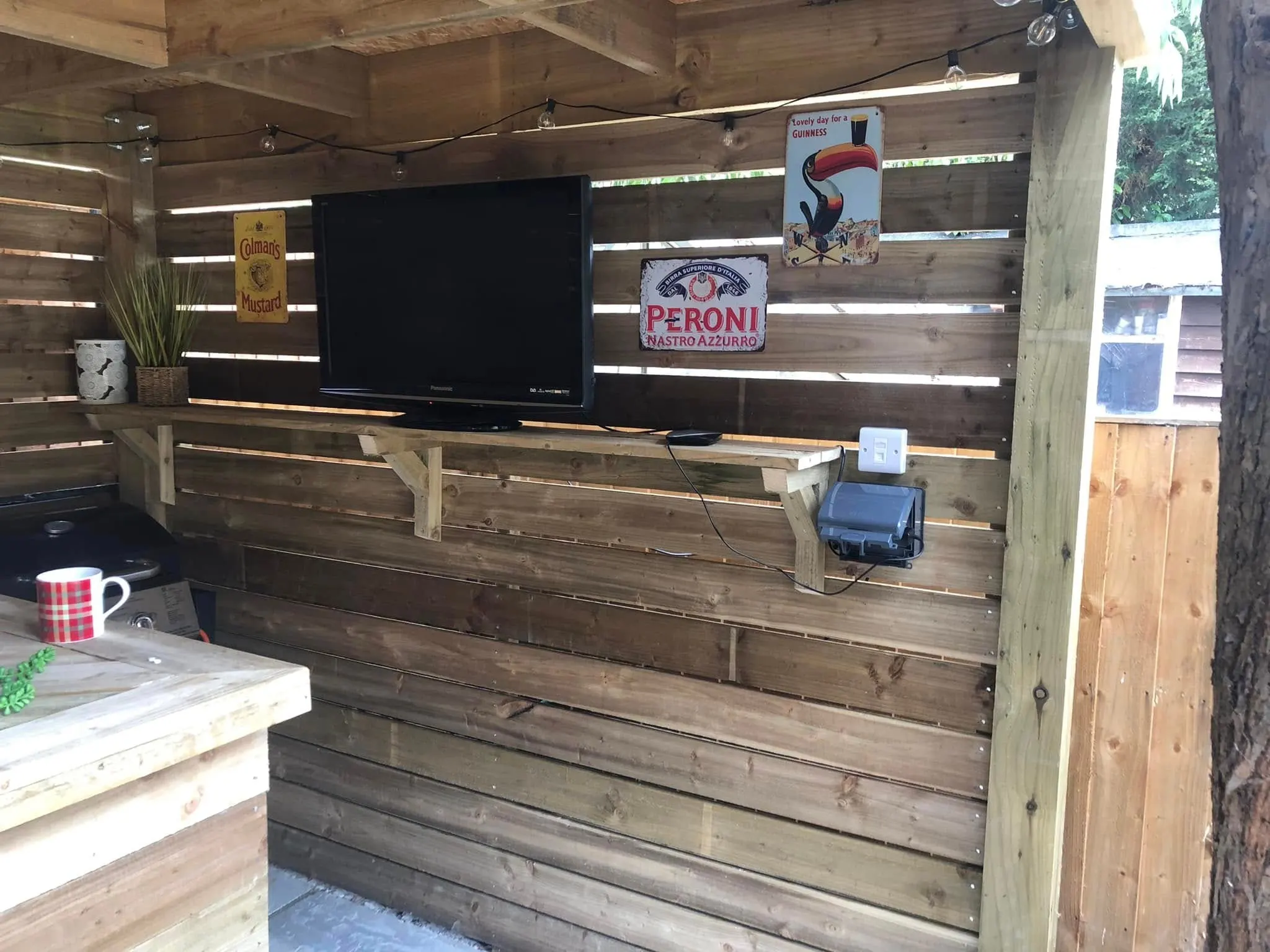 TV on a timber shelf in a slatted garden bar, with a white data outlet and an outdoor power socket beside it