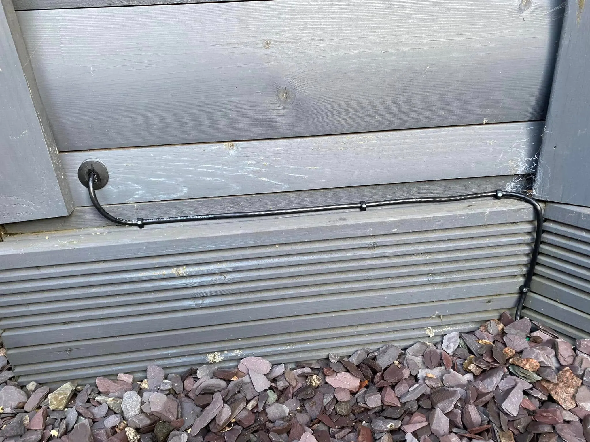 Black network cable clipped along grey cabin boards and entering the wall through a rubber grommet