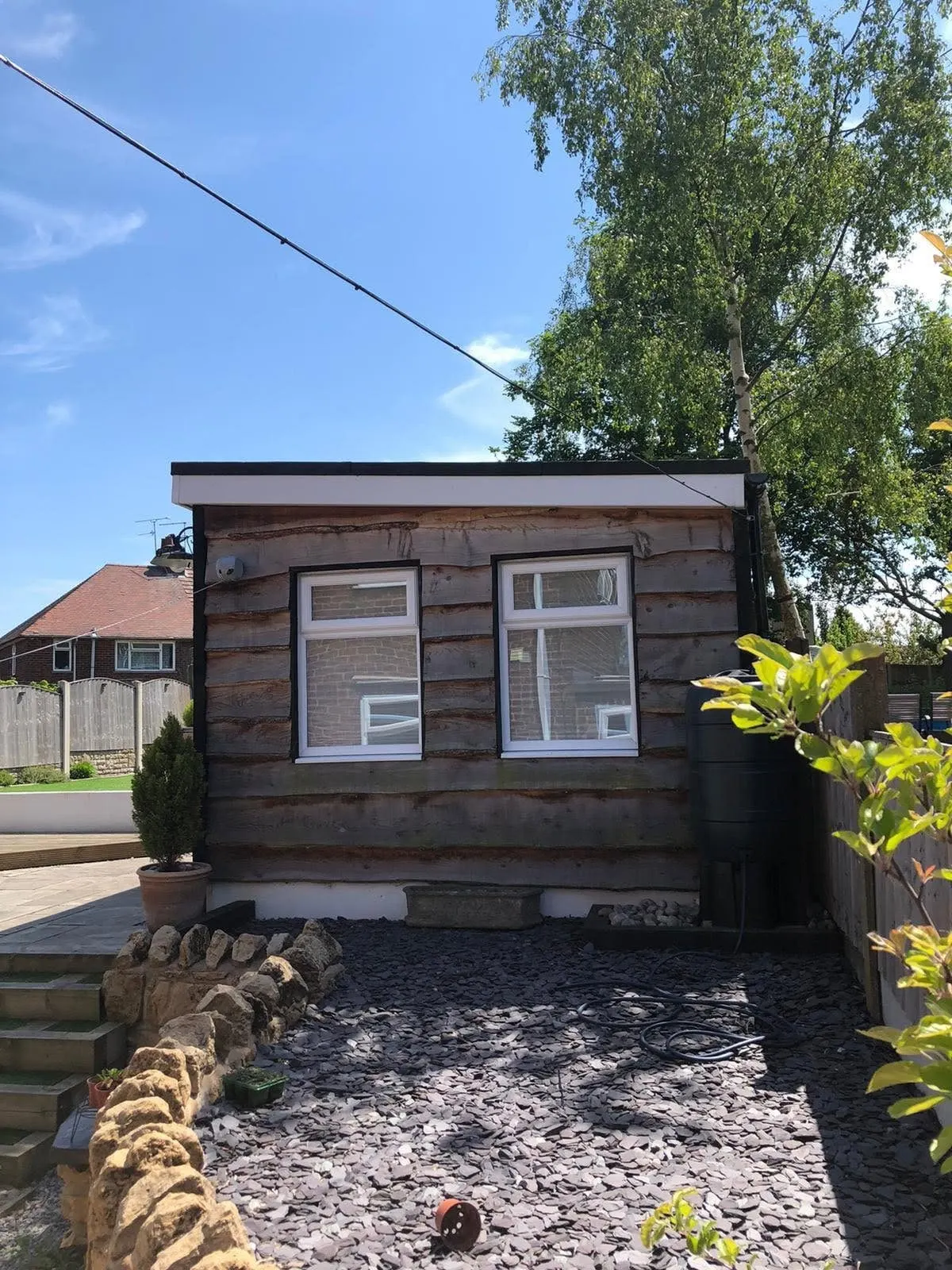 End view of a waney-edge garden office with a black cable on a catenary wire crossing the sky to its corner