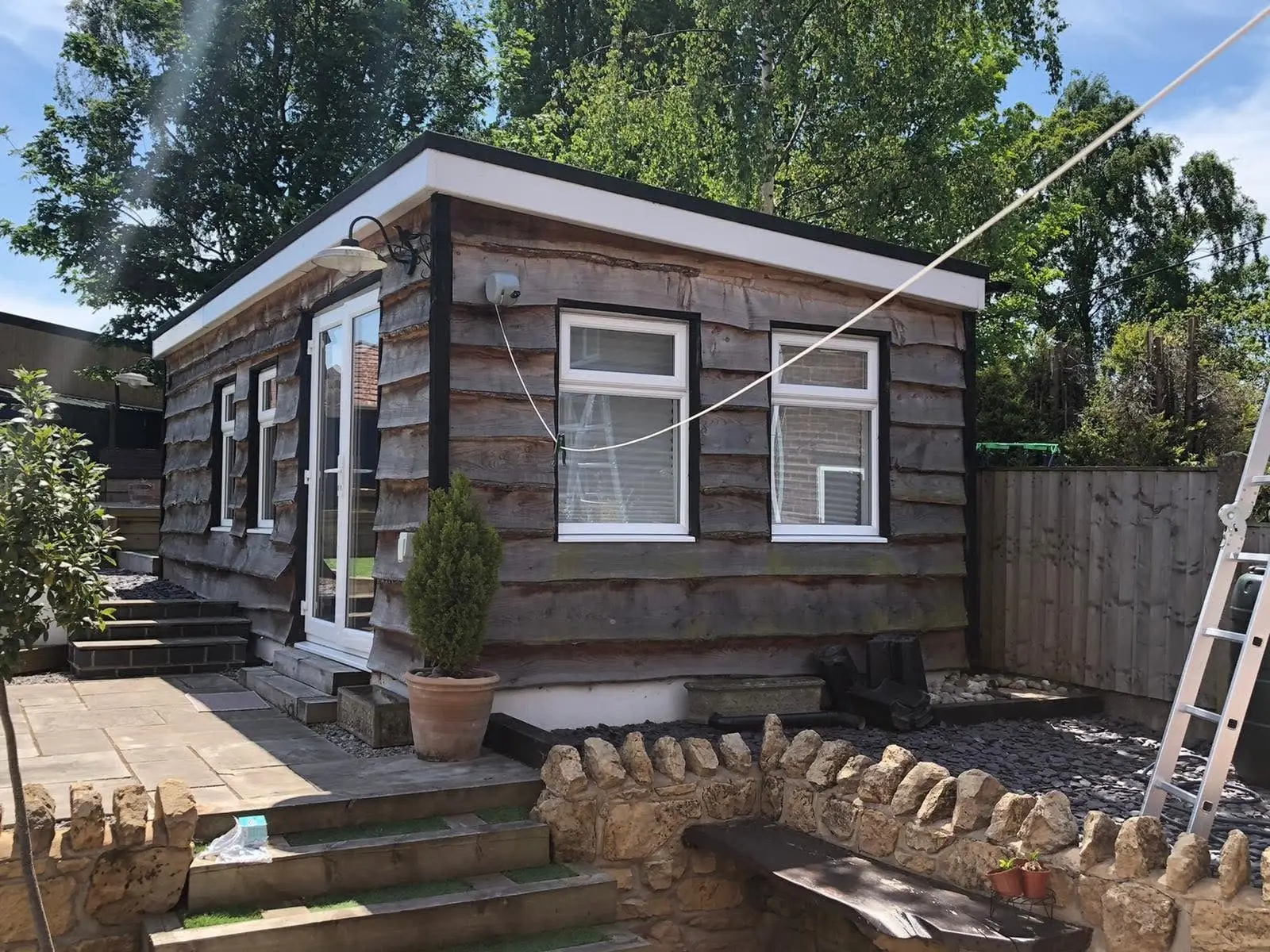 Waney-edge timber garden office with a white cable on a catenary wire running across the garden to its front wall