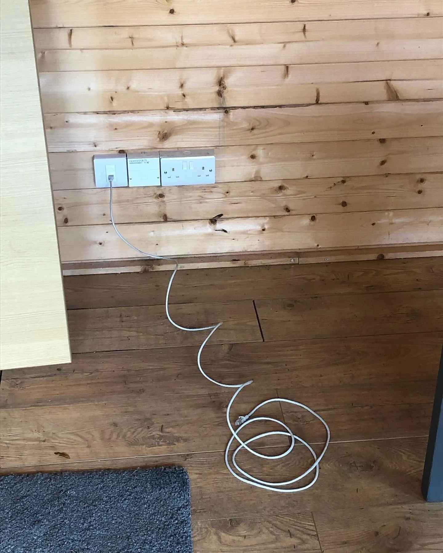 White data outlet beside an Openreach socket and double power socket on a pine-clad wall, with a patch lead to the floor
