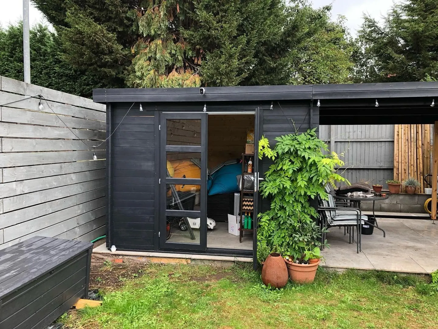 Black-stained timber garden room with festoon lights, an open glazed door and a covered patio alongside