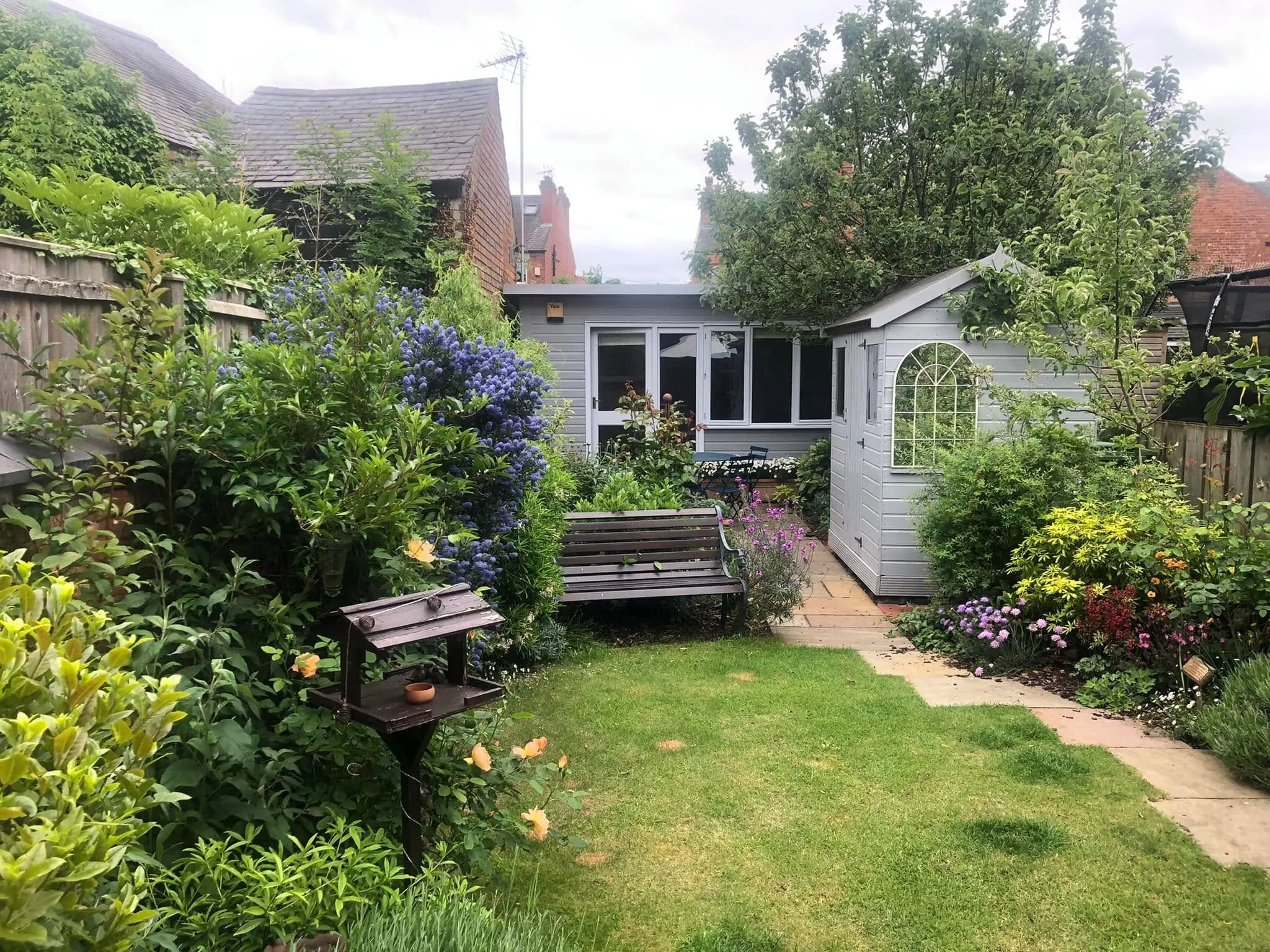 Pale grey summerhouse office and matching shed at the end of a flowering cottage garden with a bench and lawn