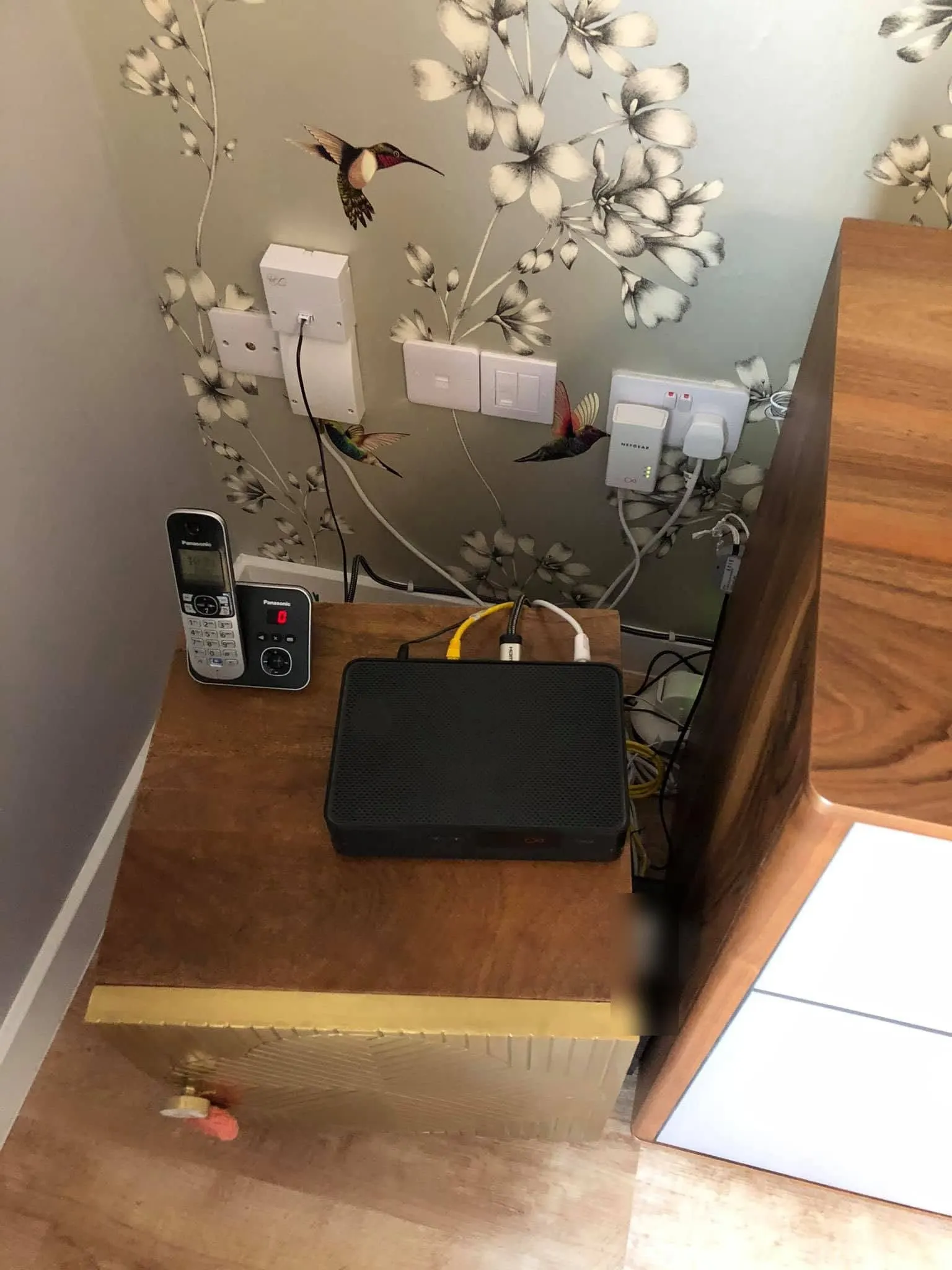 Black Virgin set-top box with HDMI and ethernet leads on a wooden bedside cabinet, below wall faceplates and a Netgear powerline adapter
