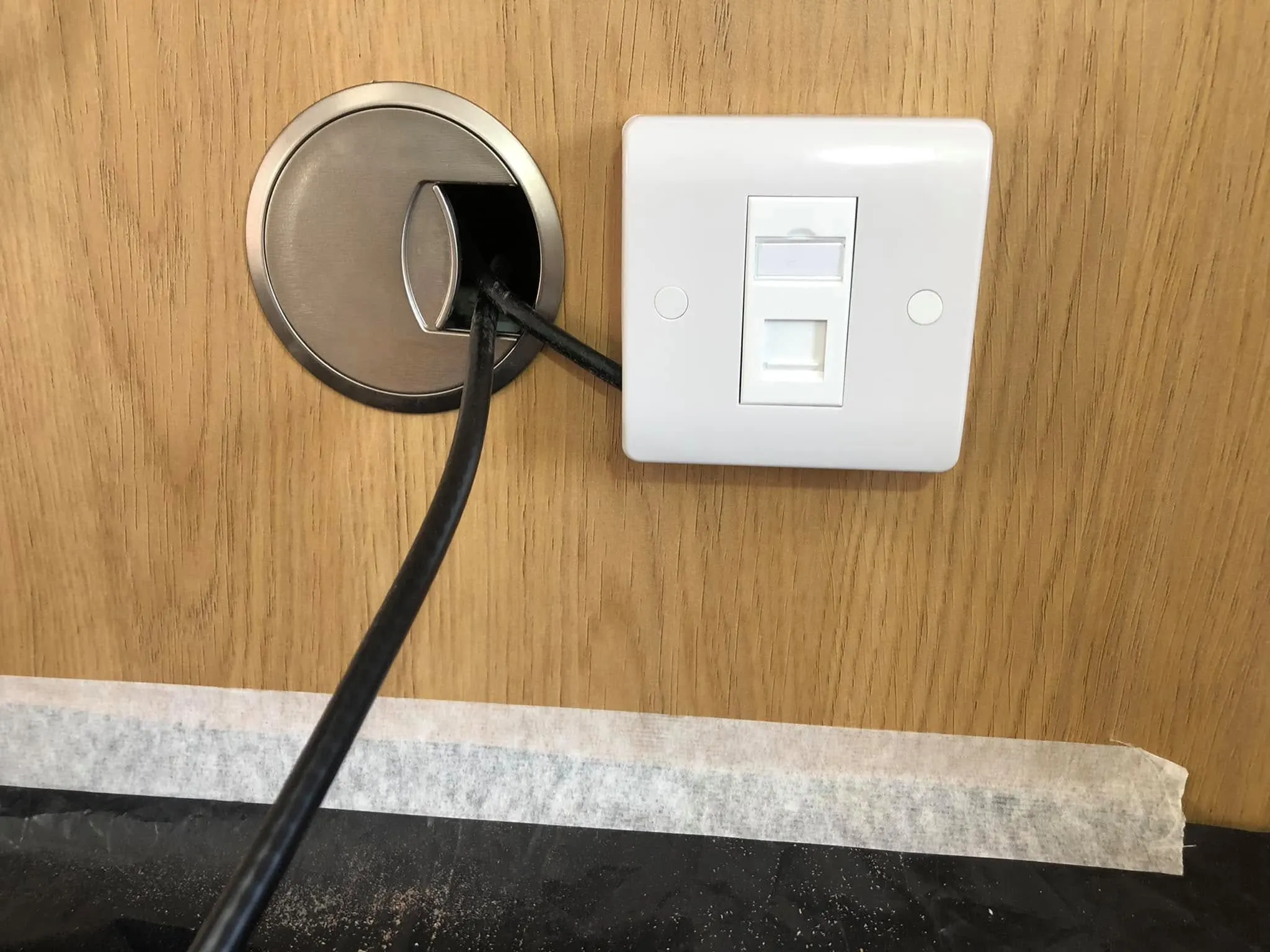 Single ethernet faceplate on an oak panel beside a brushed steel desk grommet with black cables running through