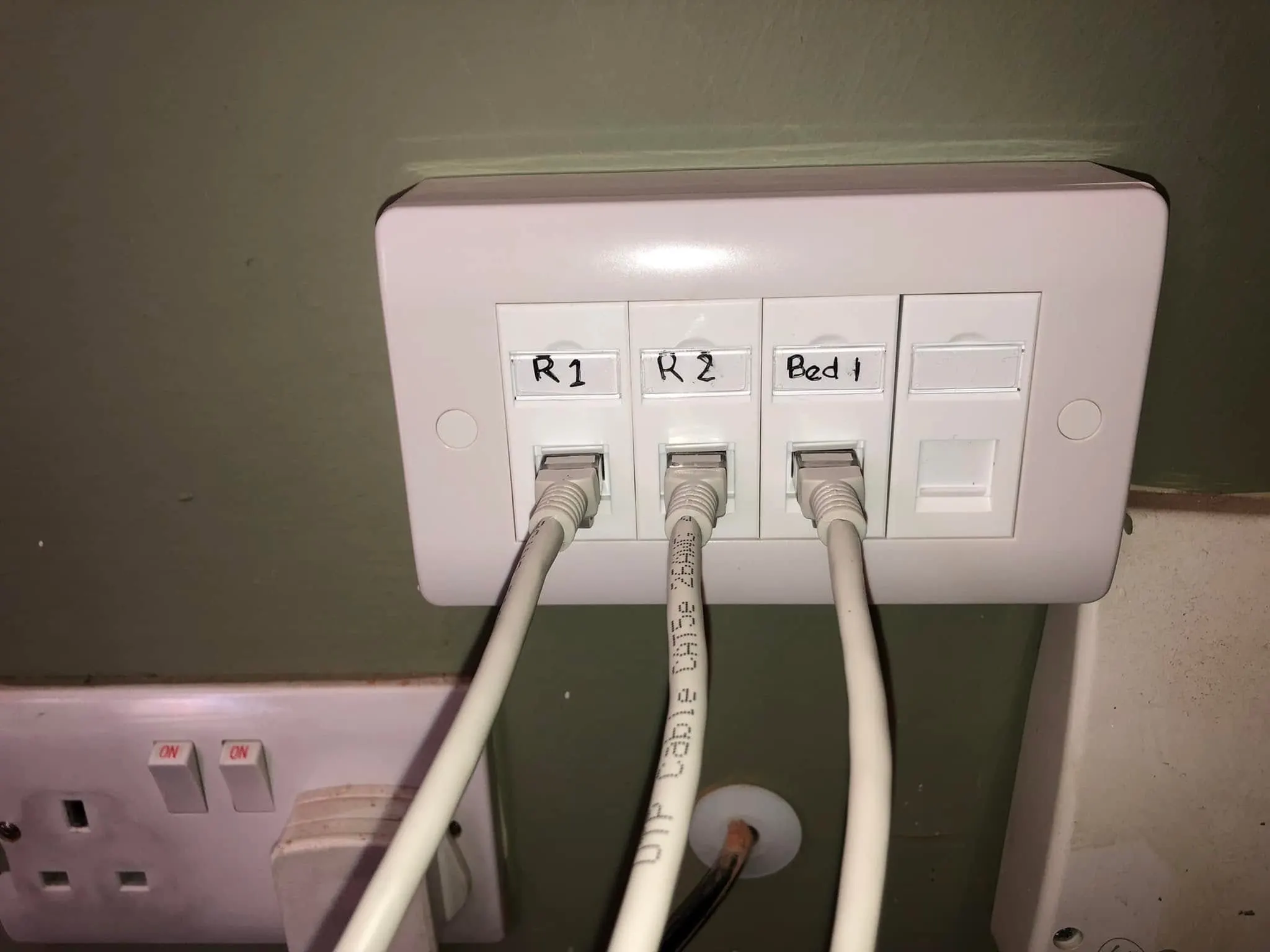 Four-port ethernet faceplate labelled R1, R2 and Bed 1 with grey patch leads plugged in, on a green wall