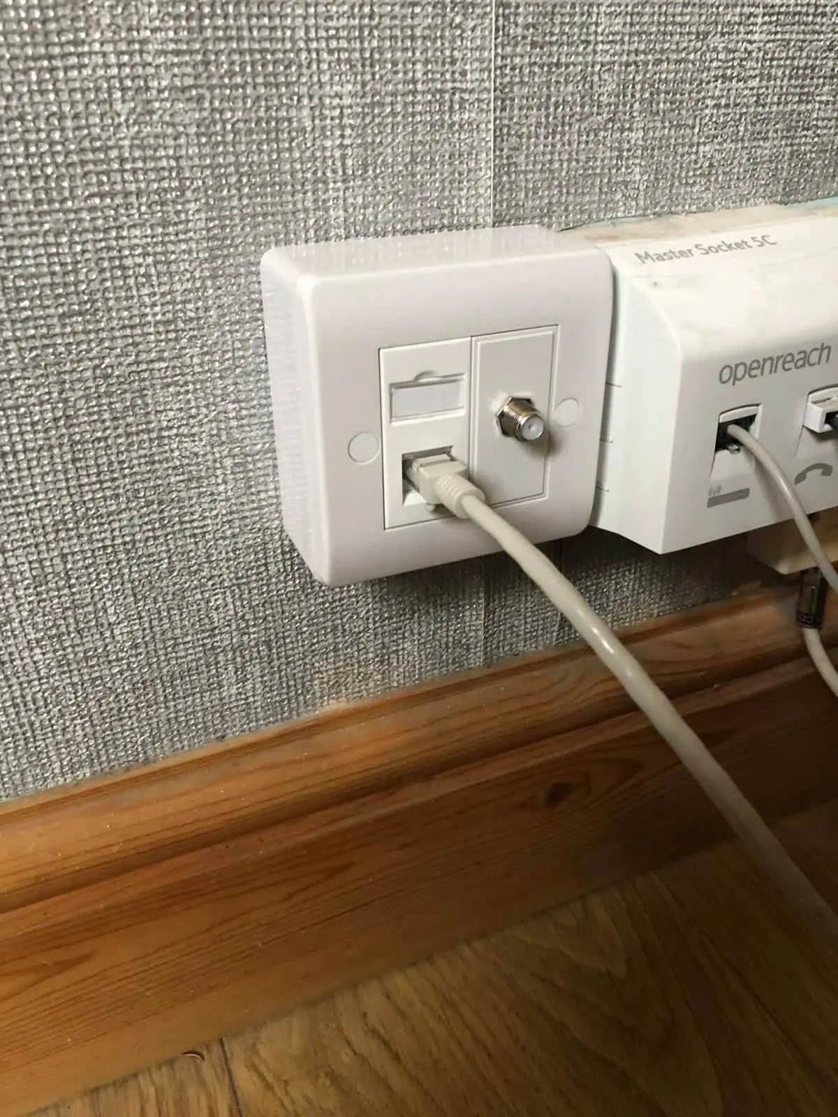 Faceplate with an ethernet port and an F-type coax connector, beside an Openreach master socket on grey textured wallpaper