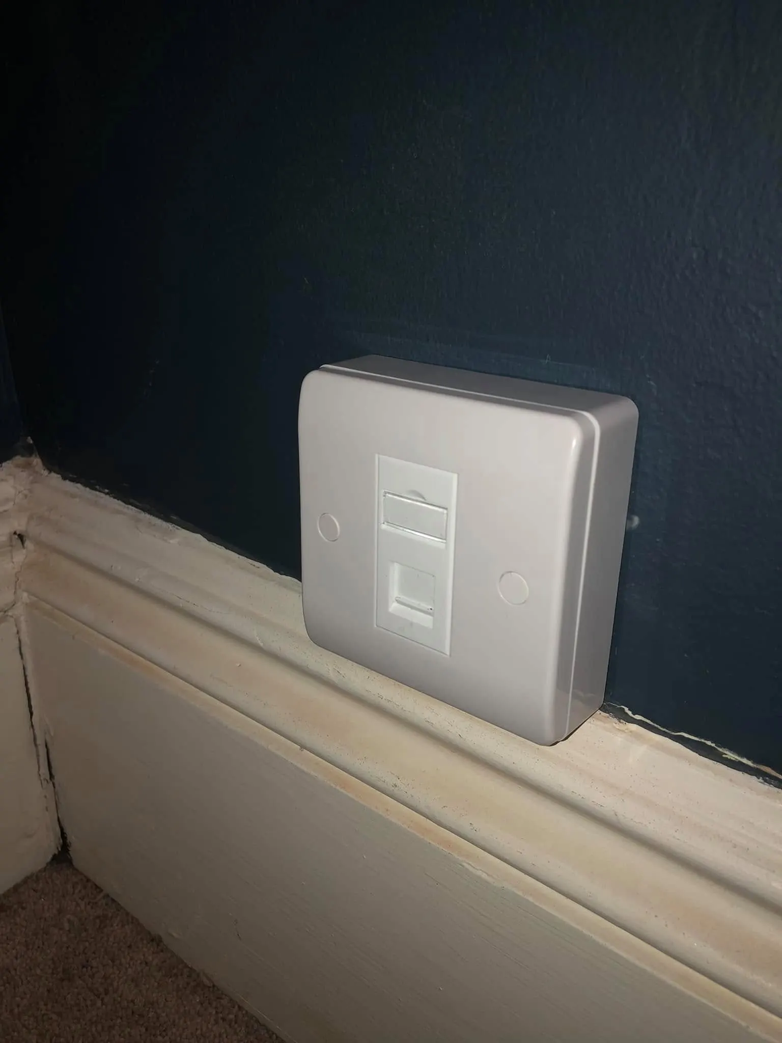Single white ethernet socket on a surface box against a dark navy wall, resting on a white skirting board