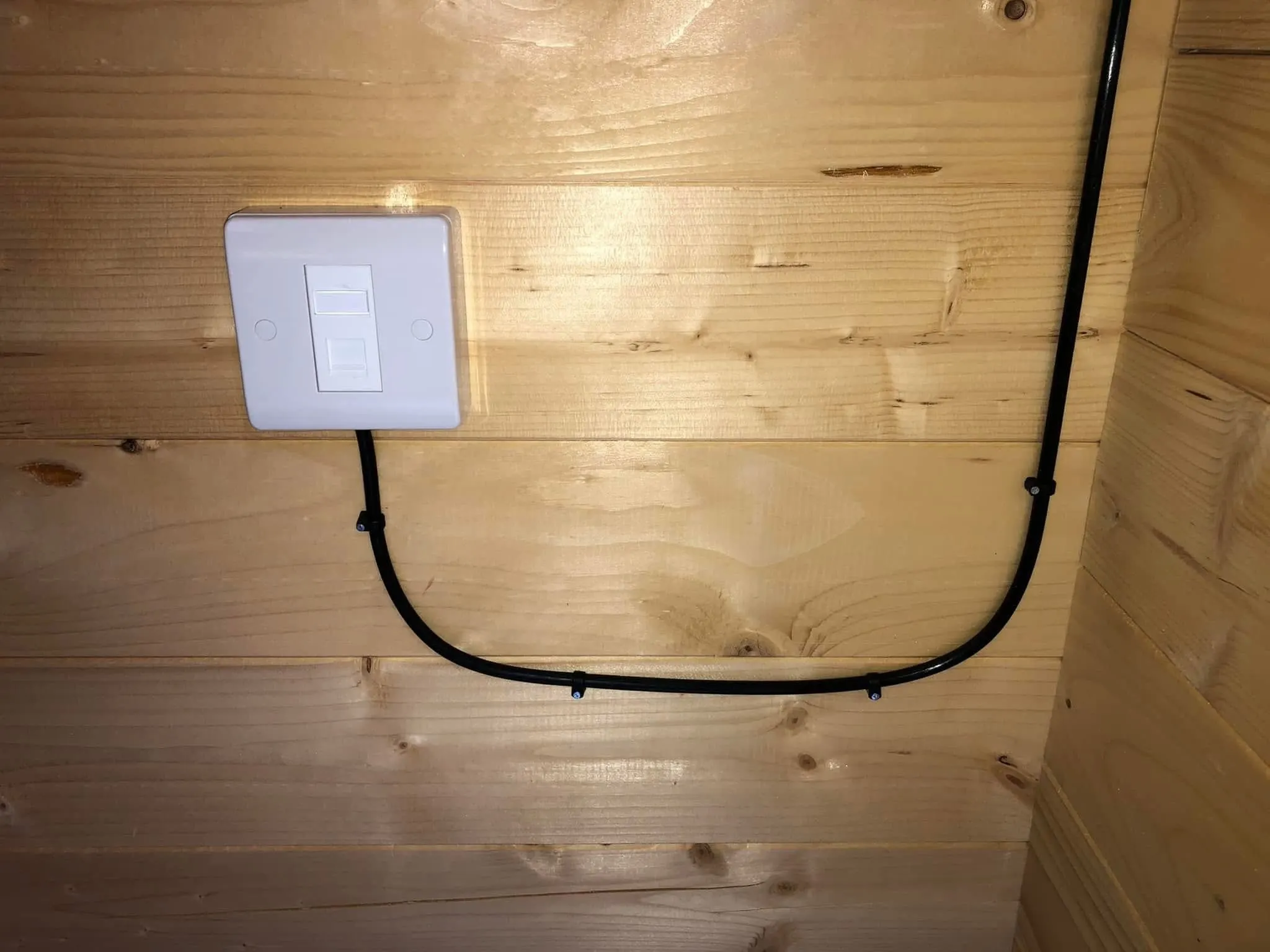 Single ethernet faceplate on pine cladding with a black cable clipped in a smooth curve up into the corner